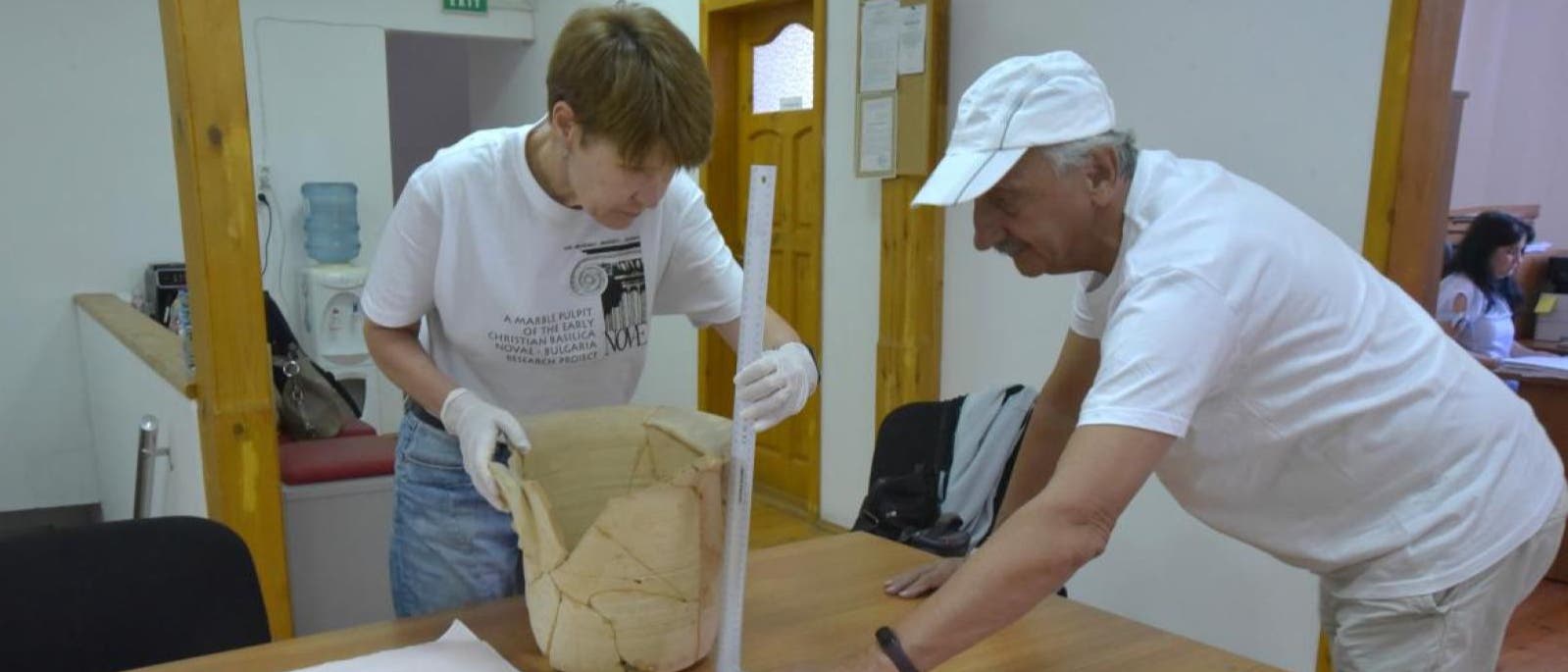 Zwei Personen in einem Raum f&uuml;hren arch&auml;ologische Arbeiten durch. Eine Person h&auml;lt ein gro&szlig;es, restauriertes Keramikgef&auml;&szlig;, w&auml;hrend die andere mit einem Ma&szlig;stab die H&ouml;he des Gef&auml;&szlig;es misst. Beide tragen wei&szlig;e T-Shirts und die Person, die das Gef&auml;&szlig; h&auml;lt, tr&auml;gt Handschuhe. Im Hintergrund sind ein Wasserspender und eine T&uuml;r zu sehen.