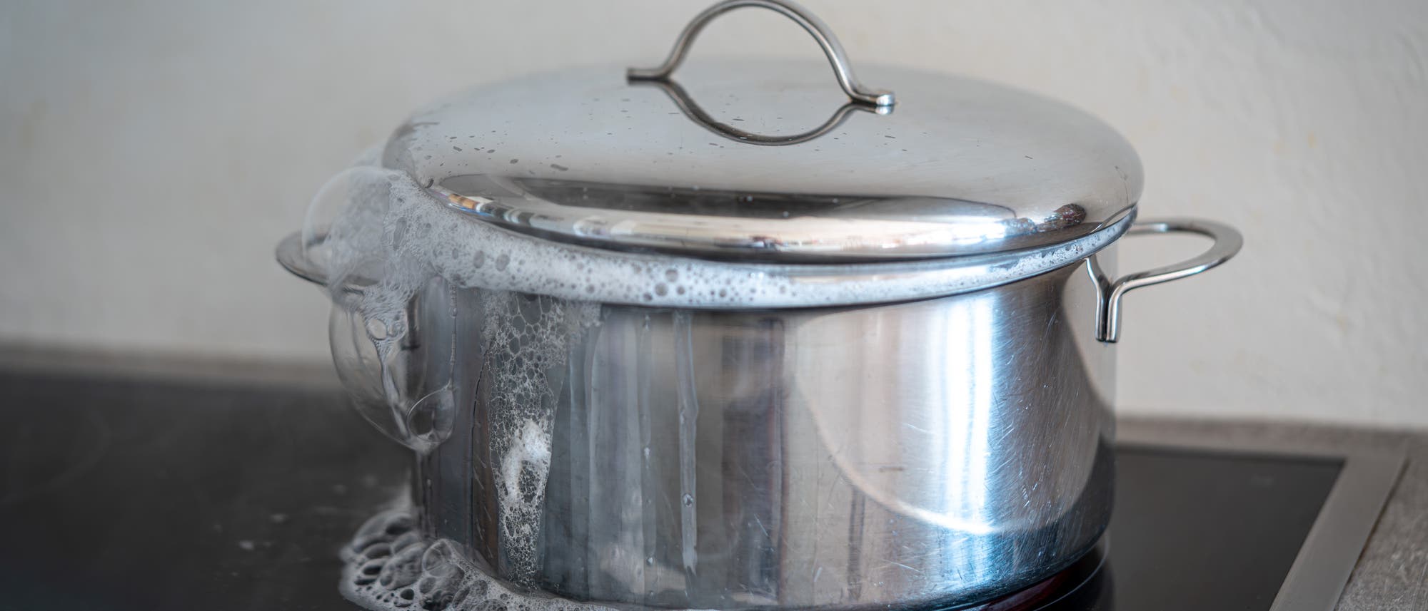 Ein Topf mit Deckel steht auf einem Herd. Schaum und Wasser laufen &uuml;ber den Rand des Topfes und verteilen sich auf der Herdplatte. Der Topf ist aus Edelstahl und spiegelt das Licht. Die Szene zeigt eine typische K&uuml;chensituation, in der etwas &uuml;berkocht.