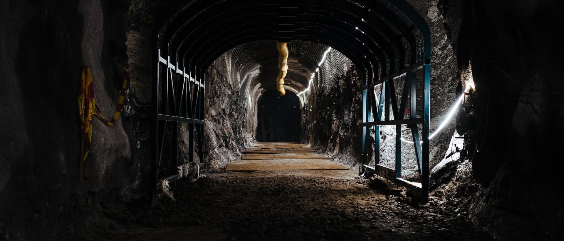 Ein beleuchteter Tunnel mit stützenden Metallbögen und einer unebenen, erdigen Oberfläche. An den Wänden sind einige farbige Markierungen zu sehen. Der Tunnel verläuft geradeaus und ist von dunklen, felsigen Wänden umgeben. Oben verläuft ein Rohr entlang der Decke.
