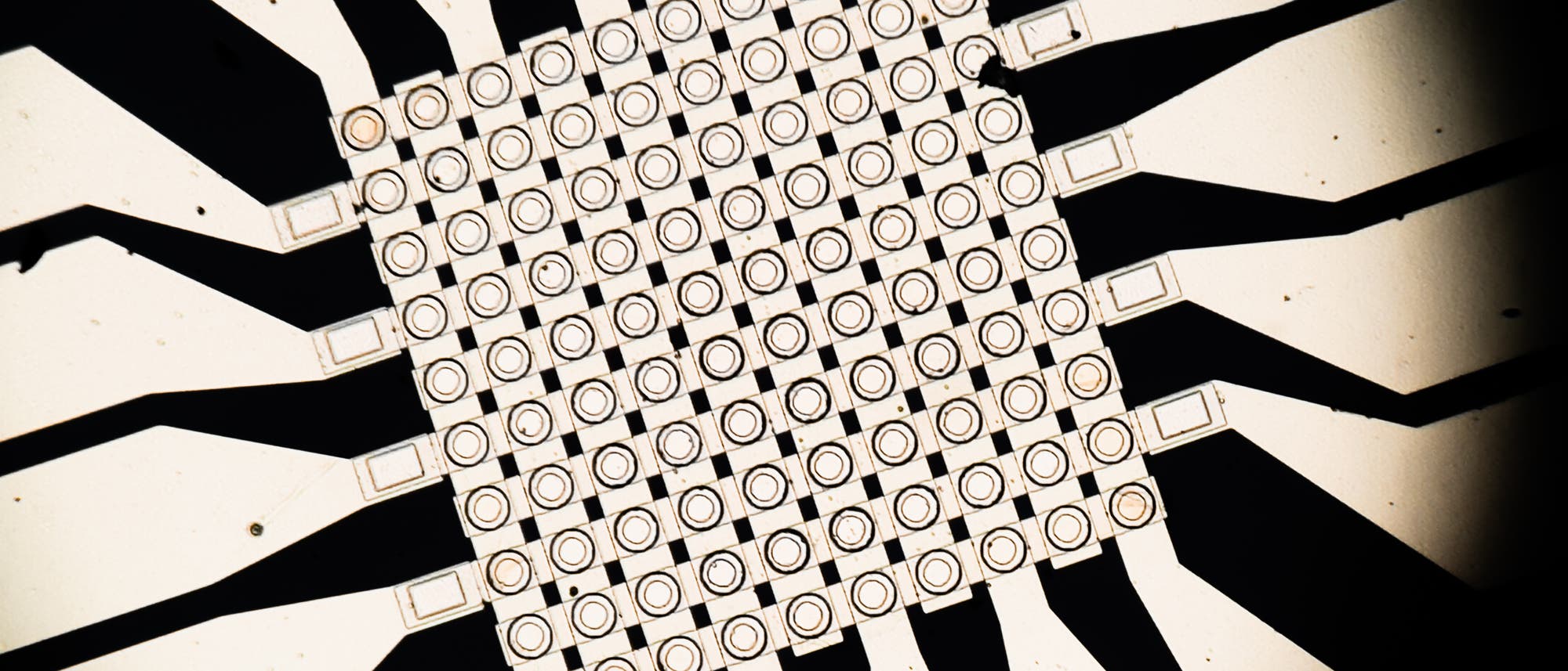 Ein Schaltkreis-Diagramm zeigt ein komplexes Gitter aus miteinander verbundenen quadratischen Elementen, die in einem symmetrischen Muster angeordnet sind. Von jedem Element führen schwarze Leiterbahnen radial nach außen. Das Bild veranschaulicht die Struktur und Verbindungspunkte eines elektronischen Schaltkreises, der für die Signalverarbeitung oder Datenübertragung verwendet werden könnte.