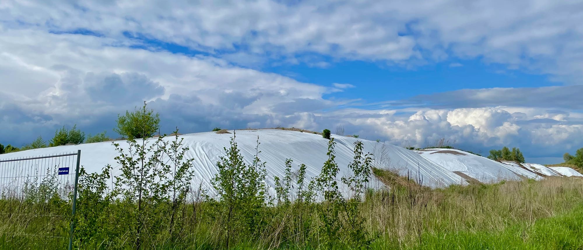 Eine weitläufige Landschaft mit grünen Wiesen und Büschen im Vordergrund. Im Hintergrund sind große, mit weißen Planen abgedeckte Hügel zu sehen, die möglicherweise landwirtschaftliche Materialien schützen. Der Himmel ist teils bewölkt mit blauen Abschnitten. Ein Zaun mit einem kleinen blauen Schild ist links im Bild sichtbar.