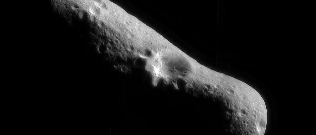 Der Asteroid Eros ist in Grau vor einem schwarzen Hintergrund zu sehen. An der Oberfläche erkennt man Krater, von denen einer in der Mitte des länglichen Körpers besonders groß ist.