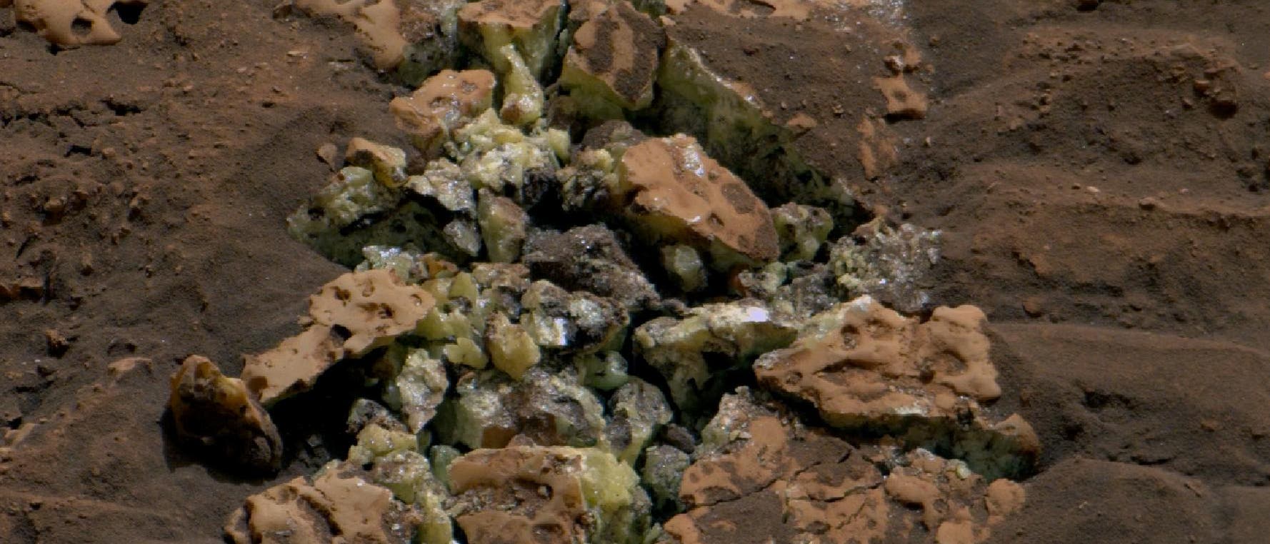 Ein zertrümmerter bräunlicher Gesteinsbrocken mit gelben Kristallen zwischen den Fragmenten.