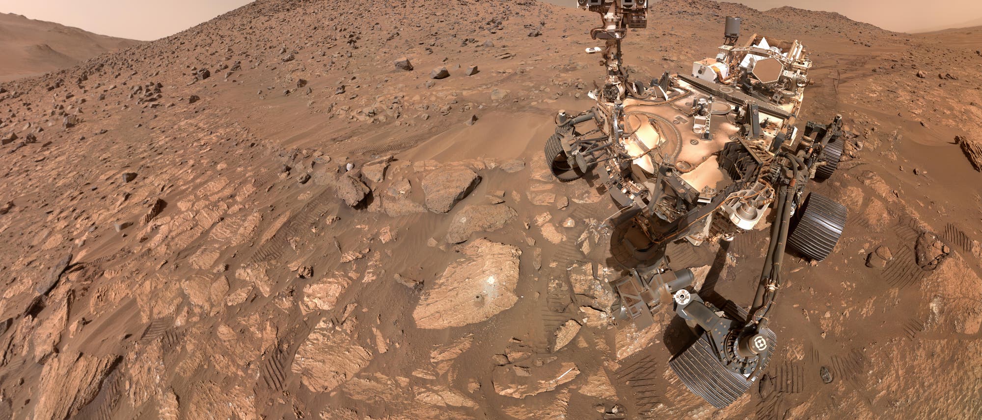 Ein Marsrover auf einer felsigen, rötlichen Marslandschaft. Der Rover ist teilweise sichtbar, mit Kameras und Instrumenten auf einem Mast. Der Boden ist mit Steinen und Sand bedeckt, und im Hintergrund sind Hügel zu sehen. Der Himmel ist staubig und orangefarben. Ein Marsrover auf einer felsigen, rötlichen Marslandschaft. Der Rover ist teilweise sichtbar, mit Kameras und Instrumenten auf einem Mast. Der Boden ist mit Steinen und Sand bedeckt, und im Hintergrund sind Hügel zu sehen. Der Himmel ist staubig und orangefarben.