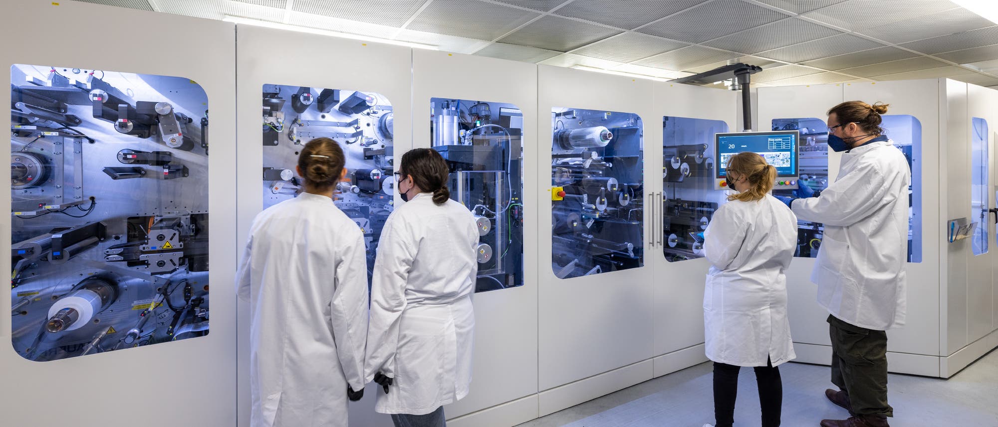 Eine Gruppe von Personen in weißen Laborkitteln steht vor einer großen, modernen Maschine in einem Labor. Die Maschine hat mehrere Fenster, durch die komplexe mechanische Teile sichtbar sind. Eine Person bedient ein Touchscreen-Display an der Maschine. Die Szene vermittelt eine professionelle, wissenschaftliche Umgebung.