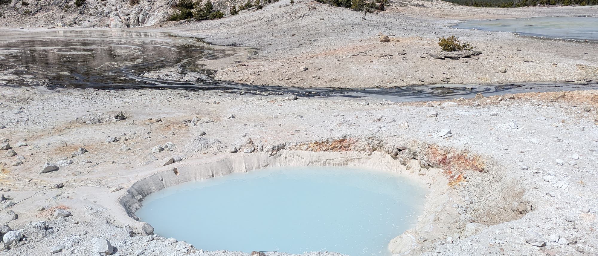 Eine bläulich-weiße heiße Quelle in einer kargen, vulkanischen Landschaft mit spärlicher Vegetation im Hintergrund. Der Himmel ist klar und blau. Die Umgebung ist steinig und mineralhaltig, typisch für geothermische Gebiete. Im Hintergrund sind einige Bäume zu sehen. Eine bläulich-weiße heiße Quelle in einer kargen, vulkanischen Landschaft mit spärlicher Vegetation im Hintergrund. Der Himmel ist klar und blau. Die Umgebung ist steinig und mineralhaltig, typisch für geothermische Gebiete. Im Hintergrund sind einige Bäume zu sehen.