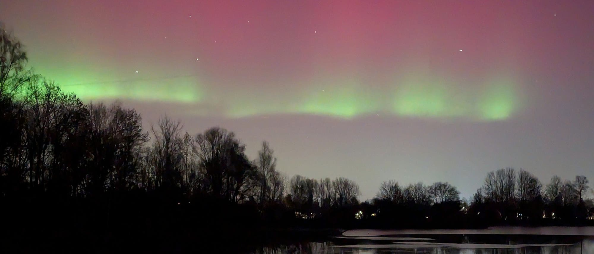 Eine n&auml;chtliche Landschaft mit einem See im Vordergrund, der die Silhouetten von B&auml;umen und das Nordlicht am Himmel reflektiert. Der Himmel zeigt gr&uuml;ne und rosa Farbt&ouml;ne, die sich &uuml;ber den Horizont erstrecken. Die Szene vermittelt eine ruhige und friedliche Atmosph&auml;re.
