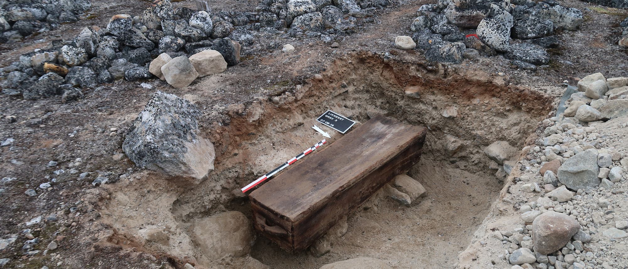 Eine archäologische Ausgrabungsstätte in einer felsigen, kargen Landschaft mit einem frei gelegten Holzsarg in einer Grube. Neben dem Sarg liegen eine Messlatte und ein kleines Schild. Im Hintergrund sind schneebedeckte Berge und ein Schiff auf dem Wasser zu sehen. Eine Person steht in der Ferne und blickt auf die Szene.