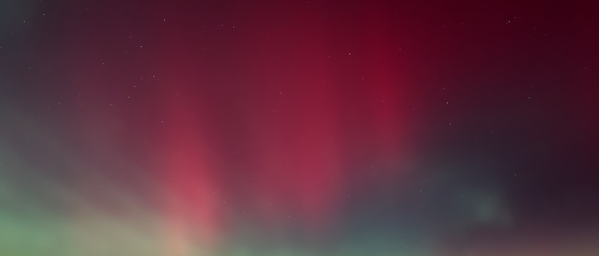 Ein nächtlicher Himmel zeigt ein beeindruckendes Polarlicht mit leuchtenden roten und grünen Farbtönen, die sich über den Horizont erstrecken. Im Vordergrund sind schwach beleuchtete Strukturen einer Stadt zu erkennen. Der Sternenhimmel ist klar sichtbar und ergänzt die natürliche Lichtshow. Ein nächtlicher Himmel zeigt ein beeindruckendes Polarlicht mit leuchtenden roten und grünen Farbtönen, die sich über den Horizont erstrecken. Im Vordergrund sind schwach beleuchtete Strukturen einer Stadt zu erkennen. Der Sternenhimmel ist klar sichtbar und ergänzt die natürliche Lichtshow.