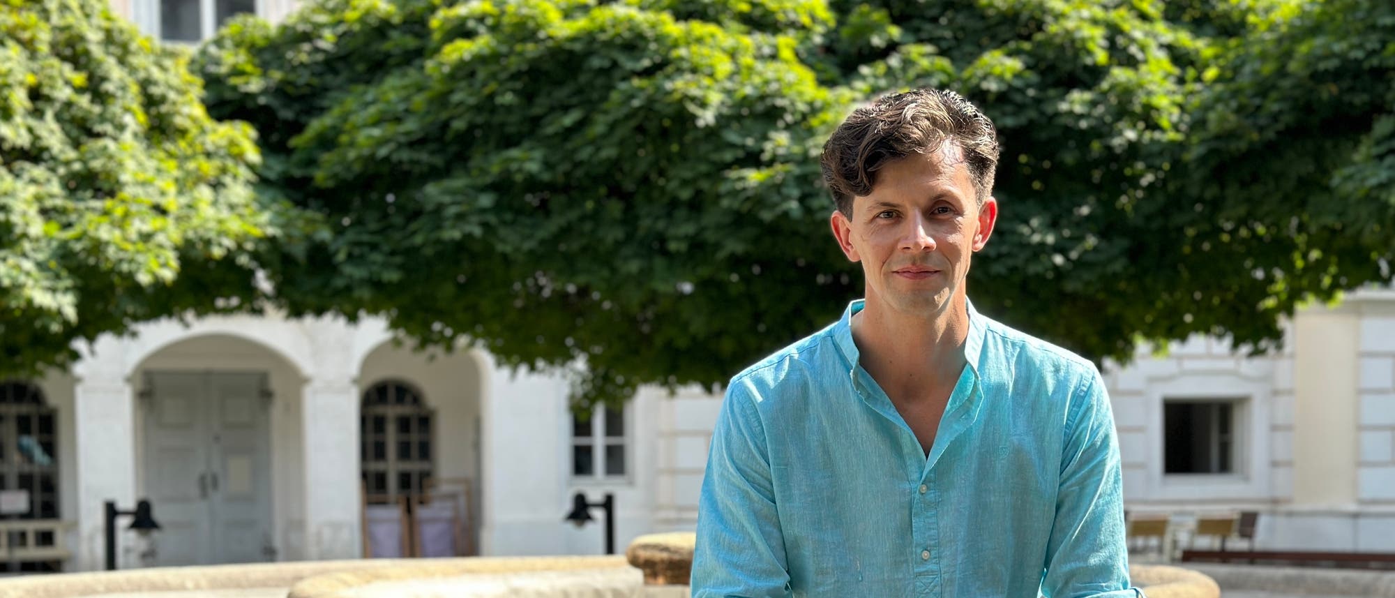 Eine Person sitzt im Freien vor einem historischen Gebäude mit weißen Wänden und großen Fenstern. Im Hintergrund sind üppige grüne Bäume zu sehen. Die Person trägt ein hellblaues Hemd und schaut in die Kamera. Die Szene wirkt ruhig und sonnig. Eine Person sitzt im Freien vor einem historischen Gebäude mit weißen Wänden und großen Fenstern. Im Hintergrund sind üppige grüne Bäume zu sehen. Die Person trägt ein hellblaues Hemd und schaut in die Kamera. Die Szene wirkt ruhig und sonnig.