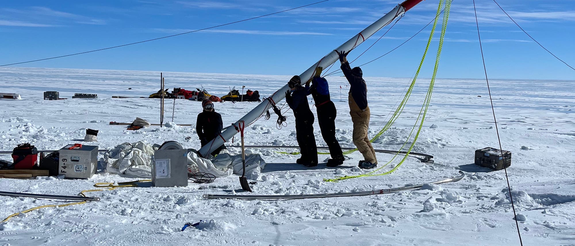 Eine Gruppe von Personen arbeitet in einer verschneiten, eisigen Landschaft der Antarktis daran, einen großen Eisbohrer aufzustellen. Der Himmel ist klar und blau. Um sie herum sind verschiedene Ausrüstungsgegenstände und Kisten auf dem Boden verteilt. Im Hintergrund sind weitere Strukturen oder Fahrzeuge zu sehen. Die Szene vermittelt den Eindruck einer wissenschaftlichen Expedition in einer abgelegenen, kalten Umgebung.