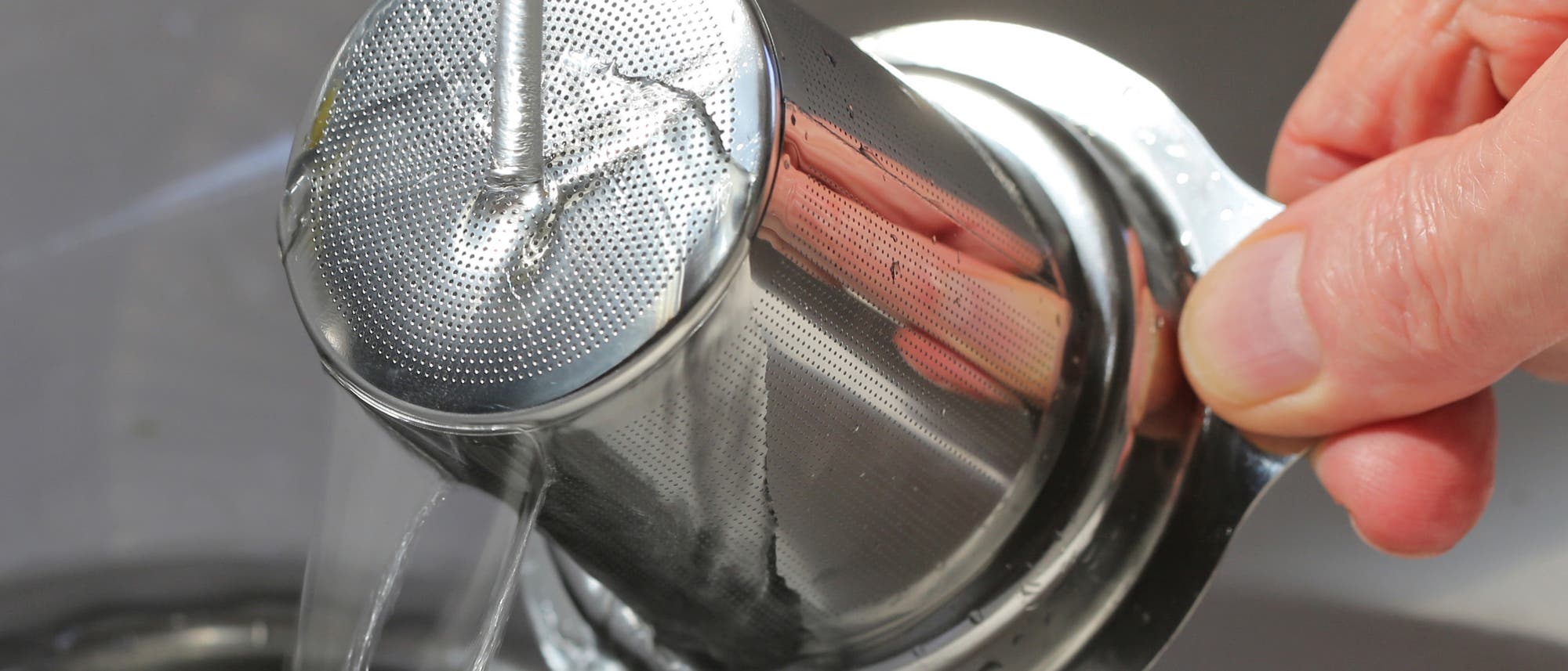 A stainless steel tea strainer is cleaned under a jet of water in the sink