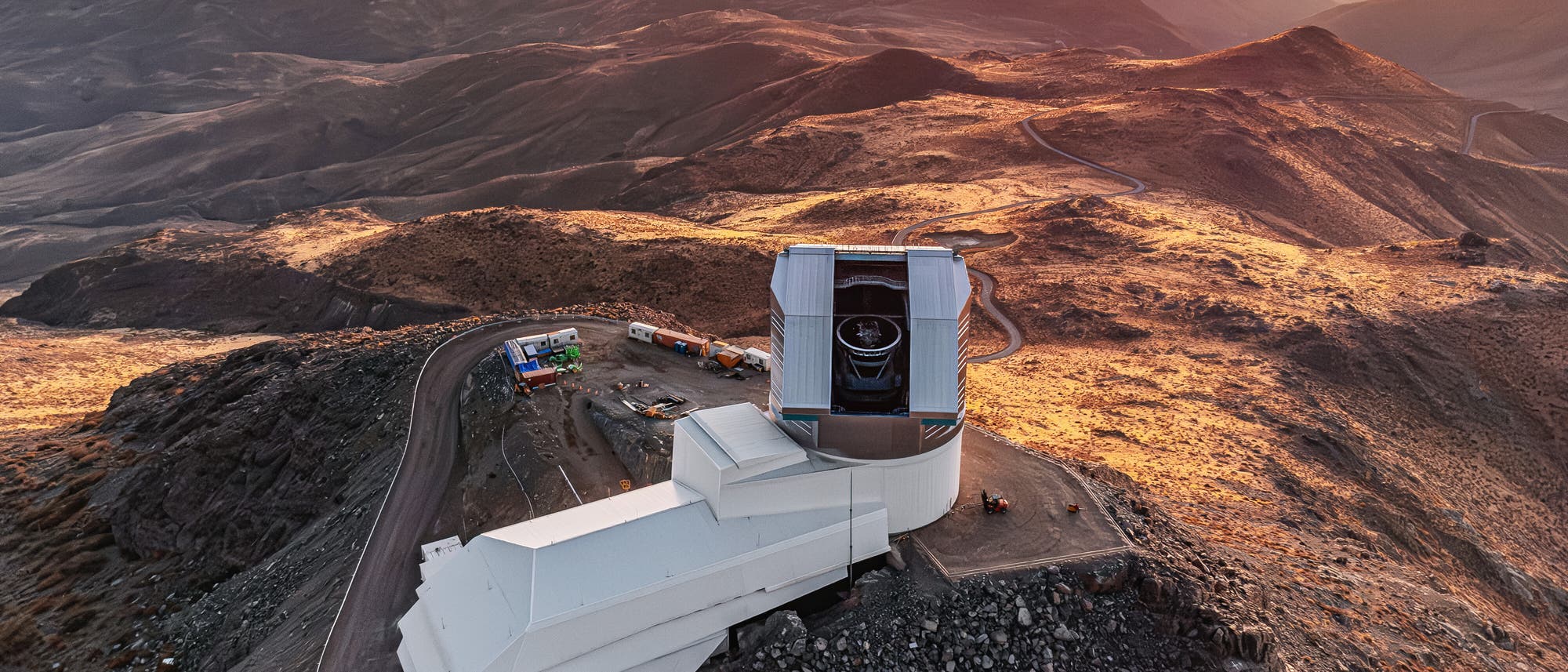 Luftaufnahme des Vera C. Rubin Observatory, das in einer bergigen W&uuml;stenlandschaft liegt. Das Geb&auml;ude hat eine moderne, wei&szlig;e Struktur mit einer gro&szlig;en Kuppel, die ein Teleskop beherbergt. Die umliegenden H&uuml;gel und T&auml;ler sind in warmes, goldenes Licht getaucht, das die raue Textur der Landschaft betont. Eine kurvenreiche Stra&szlig;e f&uuml;hrt zum Observatorium, und einige Fahrzeuge sind in der N&auml;he geparkt. Der Himmel ist klar, mit sanften Farb&uuml;berg&auml;ngen von Orange zu Blau.
