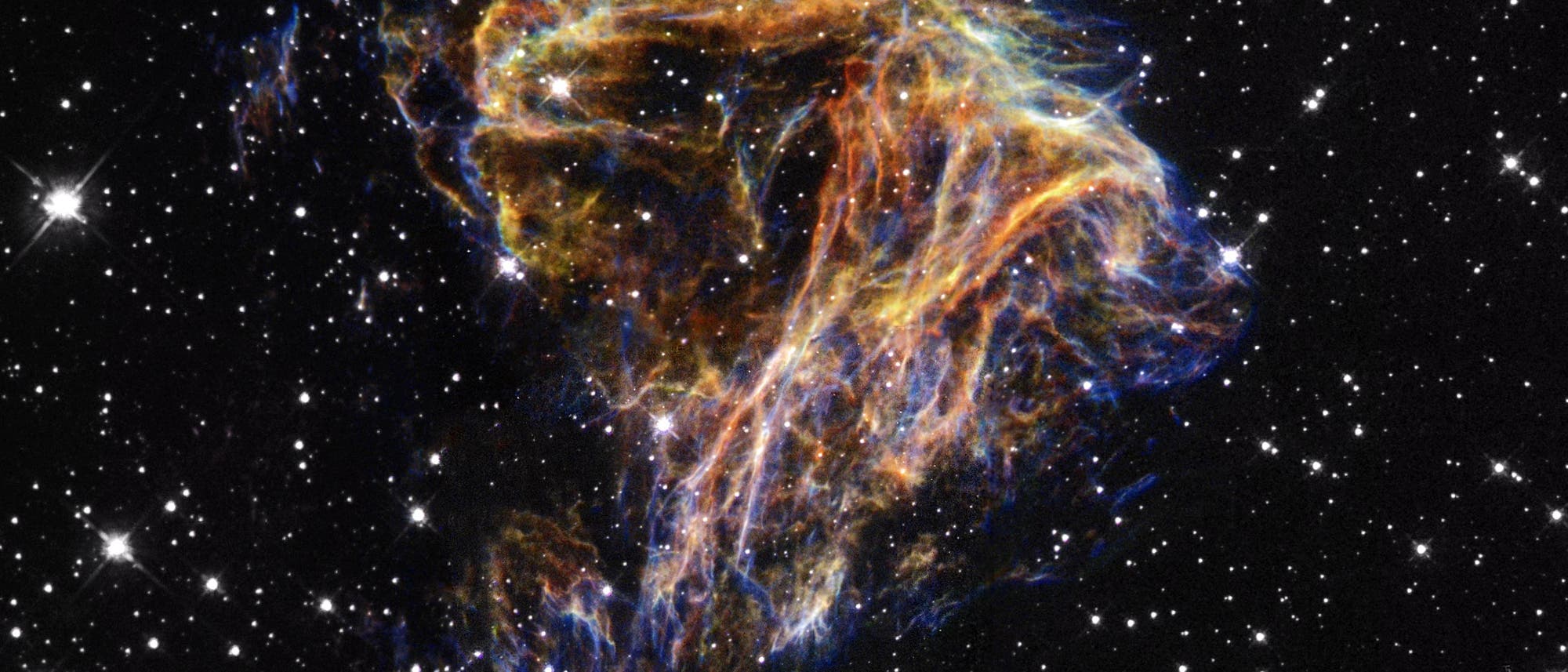 Ein farbenfrohes astronomisches Bild zeigt einen Supernova-Überrest vor einem Hintergrund aus zahlreichen leuchtenden Sternen. Die Überreste erscheinen als leuchtende, faserige Strukturen in Blau, Orange und Gelb, die sich vor dem tiefschwarzen Weltraum abheben. Die komplexen Formen und Farben illustrieren die dynamische Natur des kosmischen Ereignisses. Dieses Bild vermittelt die Schönheit und Komplexität des Universums.