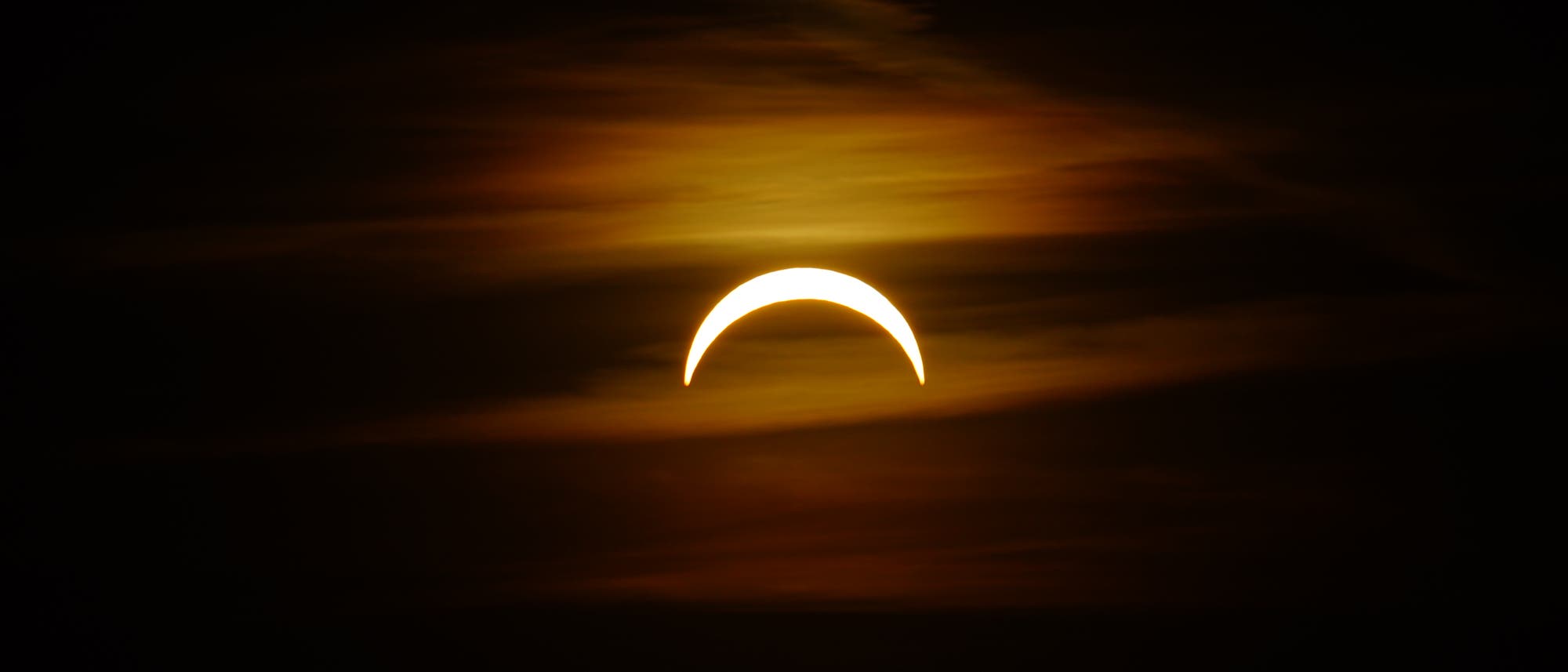 Eine partielle Sonnenfinsternis, bei der die Sonne als leuchtender, sichelf&ouml;rmiger Bogen am Himmel erscheint. Der Hintergrund ist dunkel mit einigen Wolken, die in warmen, goldenen T&ouml;nen beleuchtet sind.