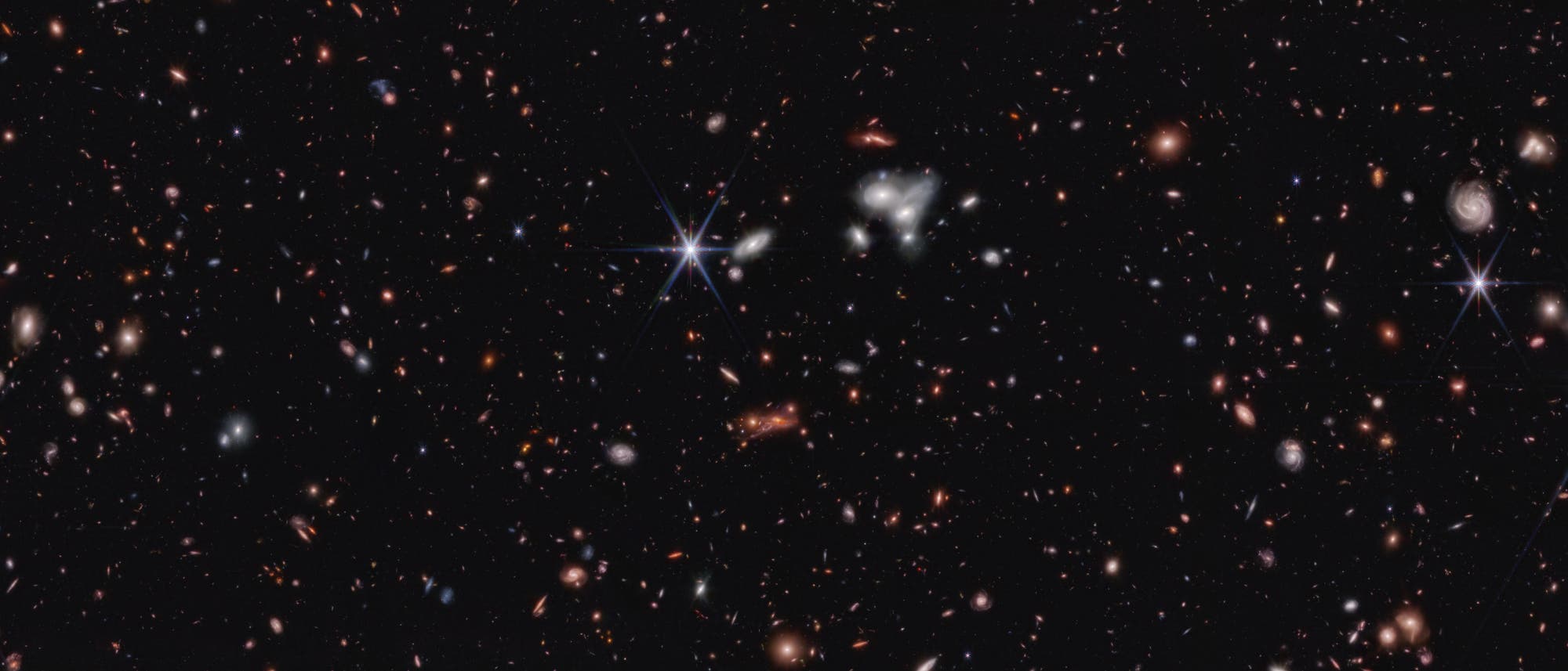 Ein tiefes Weltraumbild, das eine Vielzahl von Galaxien in unterschiedlichen Formen und Farben zeigt, die über das gesamte Bild verteilt sind. Einige Galaxien erscheinen als spiralförmige Strukturen, während andere als leuchtende Punkte oder diffuse Flecken sichtbar sind. Der Hintergrund ist schwarz, was die Helligkeit der Galaxien hervorhebt. Dieses Bild vermittelt die Weite und Vielfalt des Universums.