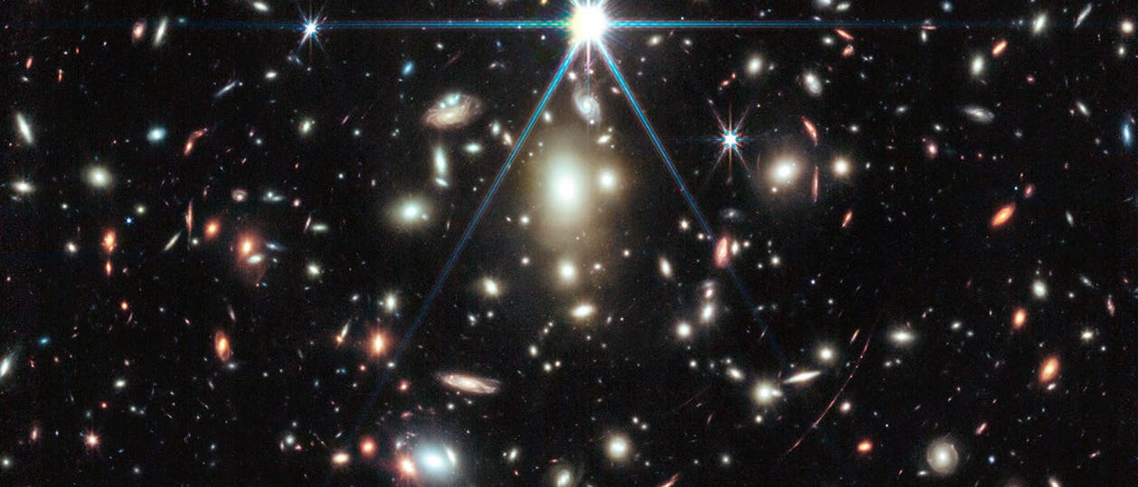 Eine Aufnahme des Weltraumteleskops, die eine Vielzahl von Galaxien in unterschiedlichen Formen und Farben zeigt, verteilt über ein dunkles, sternenübersätes Universum. Im Zentrum leuchtet ein heller Stern mit auffälligen Lichtstrahlen. Die Galaxien variieren in Größe und Helligkeit, einige erscheinen spiralförmig, andere elliptisch. Das Bild vermittelt die Weite und Vielfalt des Universums.