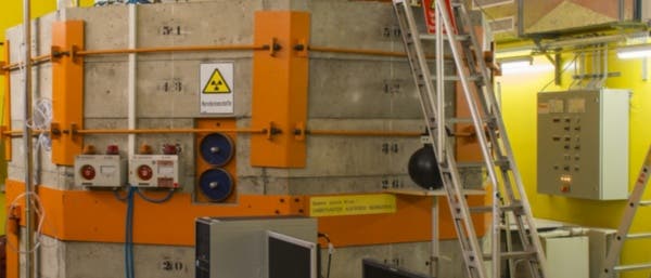 Ein Kontrollraum mit einem großen, zylindrischen Betonbehälter, der mit orangefarbenen Elementen verstärkt ist. Auf dem Behälter befindet sich ein Warnschild für Radioaktivität. Vor dem Behälter steht ein Schreibtisch mit zwei Monitoren, einer Tastatur und verschiedenen Büromaterialien. Eine Leiter führt zur Oberseite des Behälters. Im Hintergrund sind technische Geräte und Schaltkästen zu sehen. Die Umgebung wirkt industriell und funktional. Ein Kontrollraum mit einem großen, zylindrischen Betonbehälter, der mit orangefarbenen Elementen verstärkt ist. Auf dem Behälter befindet sich ein Warnschild für Radioaktivität. Vor dem Behälter steht ein Schreibtisch mit zwei Monitoren, einer Tastatur und verschiedenen Büromaterialien. Eine Leiter führt zur Oberseite des Behälters. Im Hintergrund sind technische Geräte und Schaltkästen zu sehen. Die Umgebung wirkt industriell und funktional.
