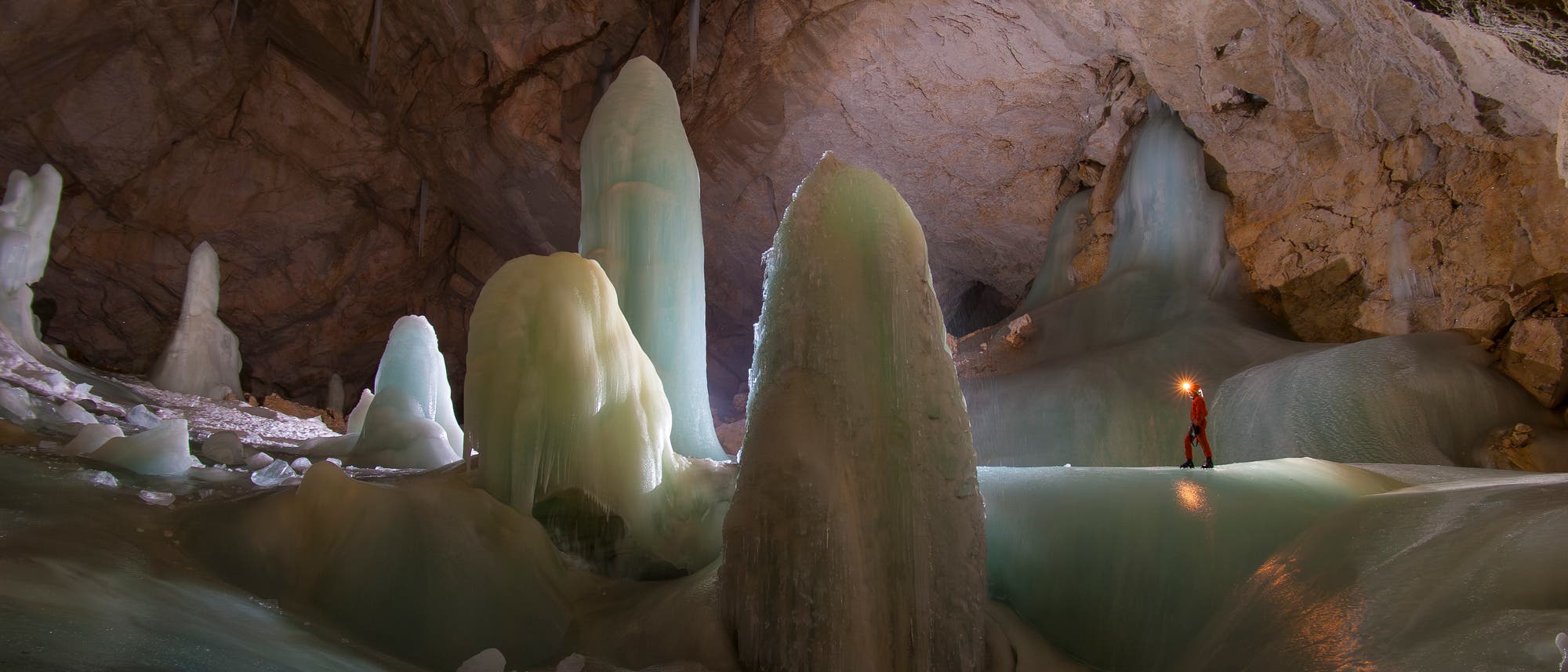 Eine Person in roter Kleidung steht mit einer Lampe in einer großen Eishöhle. Umgeben von beeindruckenden Eisformationen, die wie Stalagmiten aus dem Boden ragen. Die Höhlenwände sind aus hellem Gestein, und das Licht der Lampe reflektiert auf dem Eis, wodurch eine mystische Atmosphäre entsteht. Eine Person in roter Kleidung steht mit einer Lampe in einer großen Eishöhle. Umgeben von beeindruckenden Eisformationen, die wie Stalagmiten aus dem Boden ragen. Die Höhlenwände sind aus hellem Gestein, und das Licht der Lampe reflektiert auf dem Eis, wodurch eine mystische Atmosphäre entsteht.