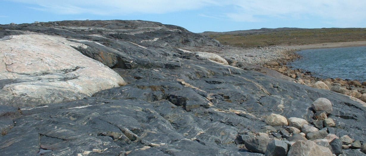 Eine felsige Küstenlandschaft mit dunklen, geschichteten Gesteinsformationen im Vordergrund. Der Himmel ist leicht bewölkt, und rechts im Bild ist ein ruhiges Gewässer zu sehen. Die Landschaft erstreckt sich in die Ferne, wobei das Ufer von kleineren Steinen gesäumt ist.
