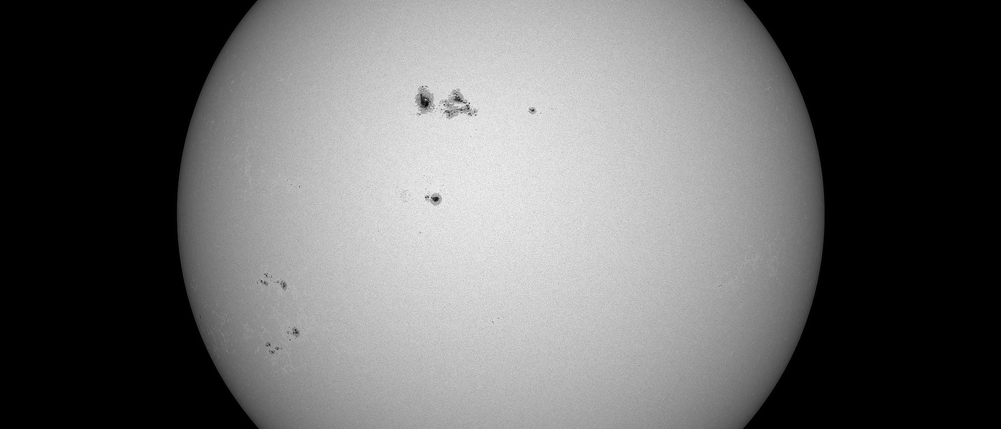 Schwarz-wei&szlig;es Bild der Sonne mit mehreren dunklen Sonnenflecken auf der Oberfl&auml;che. Die Flecken sind unregelm&auml;&szlig;ig verteilt und variieren in Gr&ouml;&szlig;e und Form. Der Hintergrund ist einheitlich schwarz, was den Kontrast zur hellen Sonnenoberfl&auml;che verst&auml;rkt. Das Bild zeigt die Sonnenaktivit&auml;t und die Struktur der Sonnenflecken.