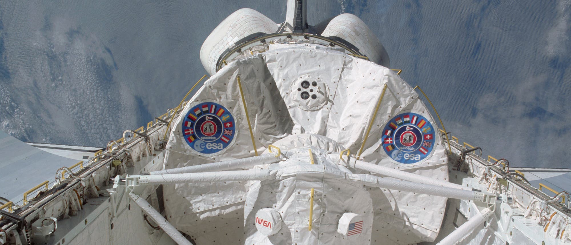 Ein Spaceshuttle im Weltraum, fotografiert von oben. Das Shuttle zeigt die Logos der NASA und ESA auf der Ladebucht. Im Hintergrund ist die Erde mit Wolkenformationen sichtbar. Die Struktur des Shuttles ist detailliert, mit sichtbaren technischen Komponenten und der US-amerikanischen Flagge.