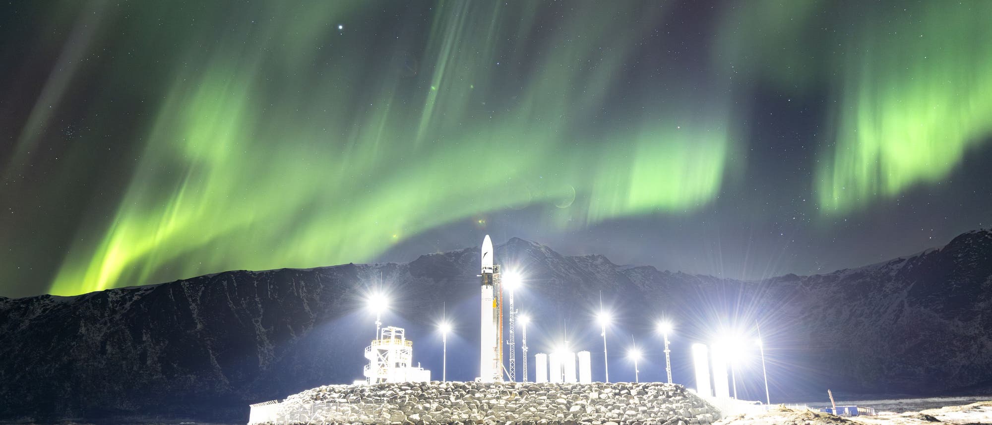 Eine Raketenabschussrampe vor einer Kulisse aus schneebedeckten Bergen und einem klaren Nachthimmel, der von grünen Nordlichtern erleuchtet wird. Helle Lichter beleuchten die Rampe und spiegeln sich im Wasser im Vordergrund wider. Der Himmel ist mit Sternen übersät, was eine ruhige und beeindruckende Atmosphäre schafft. Eine Raketenabschussrampe vor einer Kulisse aus schneebedeckten Bergen und einem klaren Nachthimmel, der von grünen Nordlichtern erleuchtet wird. Helle Lichter beleuchten die Rampe und spiegeln sich im Wasser im Vordergrund wider. Der Himmel ist mit Sternen übersät, was eine ruhige und beeindruckende Atmosphäre schafft.
