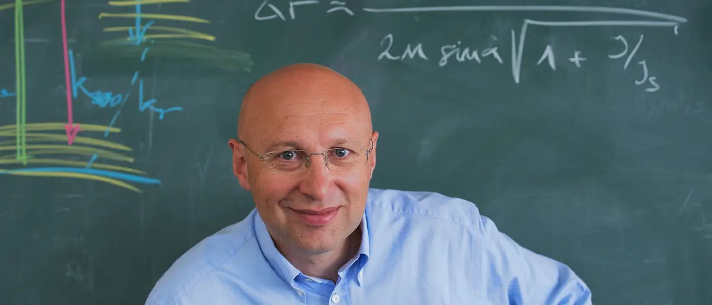 Ein Mann steht vor einer Tafel, auf der mathematische Notationen und Formeln in verschiedenen Farben geschrieben sind. Eine Formel auf der Tafel lautet: \(\Delta R = \frac{\lambda}{2m \sin \alpha} \sqrt{1 + \frac{1}{J_s}}\). Die Tafel zeigt auch farbige Diagramme und Linien. Der Mann trägt ein hellblaues Hemd und lächelt in die Kamera. Ein Mann steht vor einer Tafel, auf der mathematische Notationen und Formeln in verschiedenen Farben geschrieben sind. Eine Formel auf der Tafel lautet: \(\Delta R = \frac{\lambda}{2m \sin \alpha} \sqrt{1 + \frac{1}{J_s}}\). Die Tafel zeigt auch farbige Diagramme und Linien. Der Mann trägt ein hellblaues Hemd und lächelt in die Kamera.