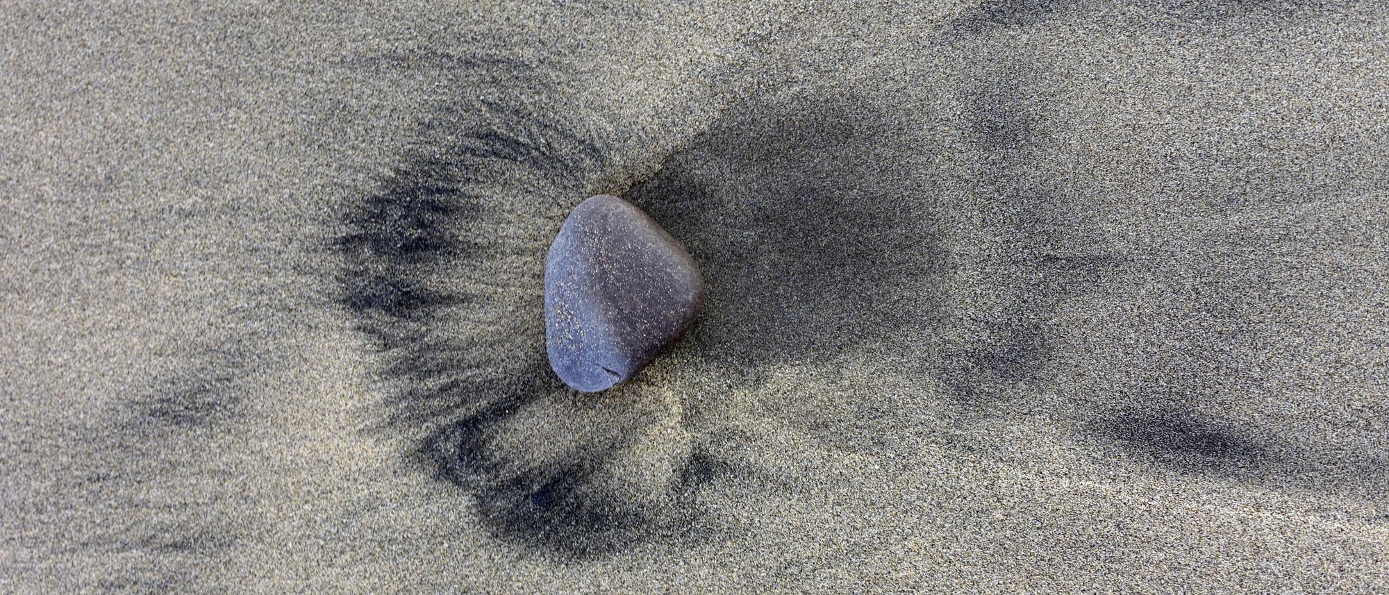 Ein einzelner grauer Stein liegt auf einer sandigen Oberfl&auml;che. Der Sand um den Stein herum zeigt ein Muster aus dunkleren und helleren Bereichen, das durch Wasserbewegungen entstanden ist. Der Stein ist leicht oval und hebt sich deutlich vom umgebenden Sand ab.