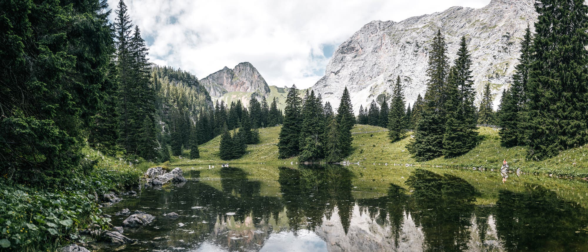 Eine malerische Berglandschaft mit einem ruhigen See im Vordergrund, der die umliegenden Tannenb&auml;ume und die felsigen Berge im Hintergrund spiegelt. Der Himmel ist teilweise bew&ouml;lkt, und das gr&uuml;ne Gras verleiht der Szene eine lebendige Farbe. Ein Wanderer ist am rechten Bildrand zu sehen, was auf Freizeitaktivit&auml;ten in der Natur hinweist.