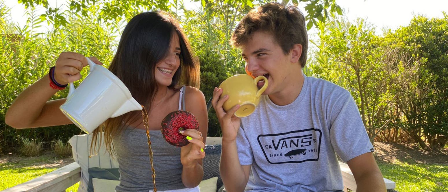 Zwei Personen sitzen im Freien auf einer Bank und lachen sich an. Die Person links hält in der rechten Hand eine Kanne und versucht in einen Donut, den sie in der rechten Hand hält, eine Flüssigkeit zu gießen. Die Person rechts hält eine Tasse und versucht hineinzubeißen. Im Hintergrund sind grüne Pflanzen zu sehen, was auf einem Garten oder Park schließen lässt. Zwei Personen sitzen im Freien auf einer Bank und lachen sich an. Die Person links hält in der rechten Hand eine Kanne und versucht in einen Donut, den sie in der rechten Hand hält, eine Flüssigkeit zu gießen. Die Person rechts hält eine Tasse und versucht hineinzubeißen. Im Hintergrund sind grüne Pflanzen zu sehen, was auf einem Garten oder Park schließen lässt.