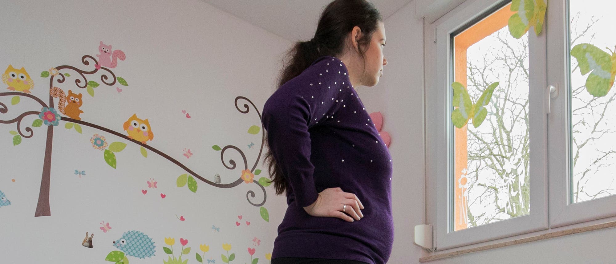 Eine schwangere Frau blickt aus einem Fenster. Sie befindet sich offenbar in einem Kinderzimmer.