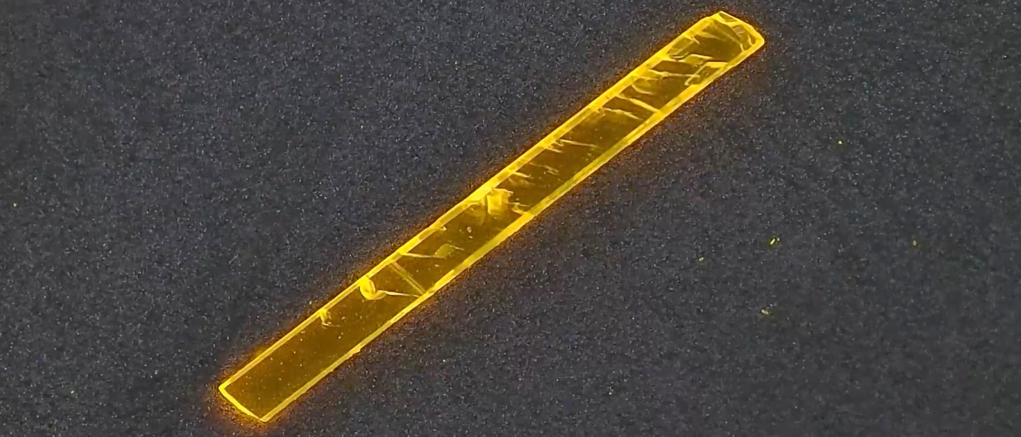 Ein fluoreszenzmikroskopisches Bild zeigt ein l&auml;ngliches, leuchtend gelbes Objekt auf einem dunklen Hintergrund. Das Objekt hat eine rechteckige Form und weist unregelm&auml;&szlig;ige Muster und Strukturen auf. Die fluoreszierende Eigenschaft hebt es deutlich vom Hintergrund ab, was auf eine spezifische Markierung oder F&auml;rbung hinweisen k&ouml;nnte. Das Bild dient der Untersuchung mikroskopischer Details und Strukturen.