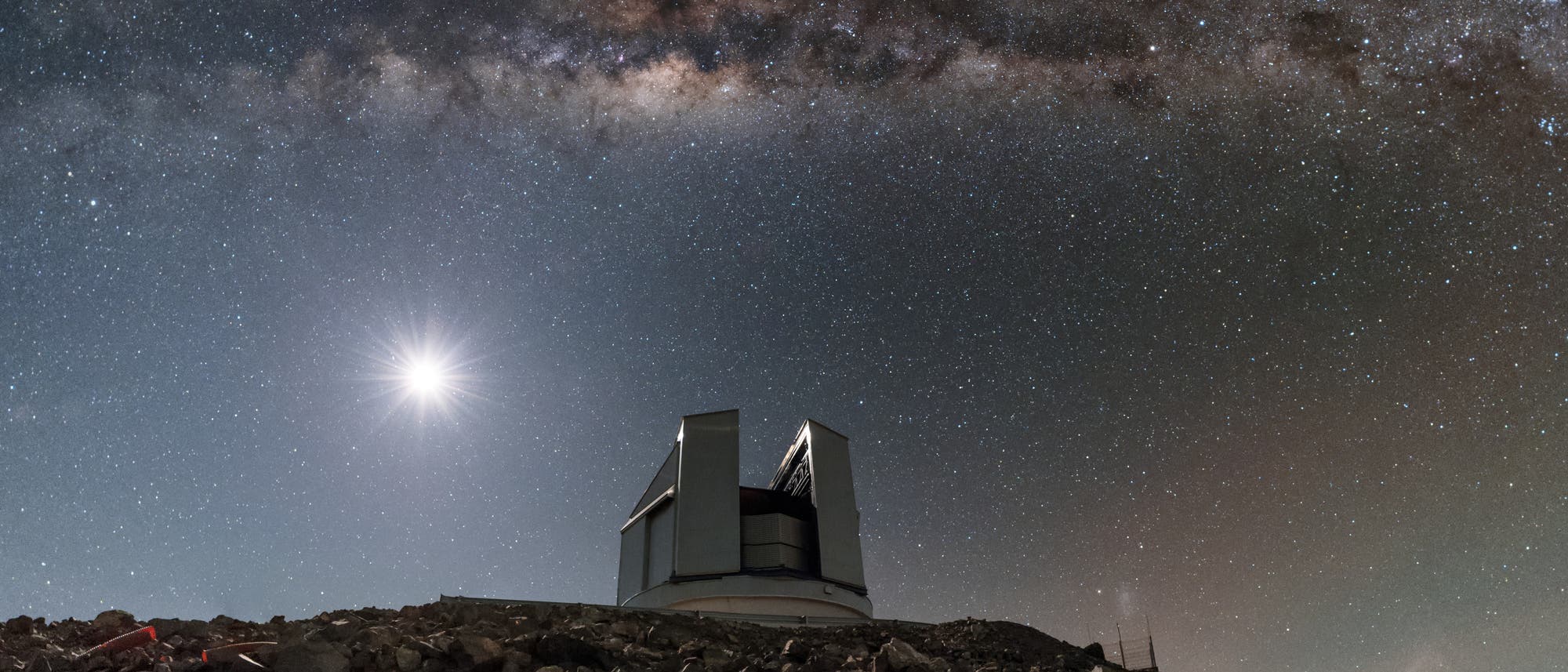 Un observatorio se encuentra en una colina rocosa bajo un cielo nocturno despejado. La Vía Láctea se extiende horizontalmente a lo largo de la imagen, mientras que un brillante cuerpo celeste, presumiblemente la Luna, brilla a la izquierda. El cielo está salpicado de numerosas estrellas, lo que constituye un impresionante telón de fondo para el observatorio. Un observatorio se encuentra en una colina rocosa bajo un cielo nocturno despejado. La Vía Láctea se extiende horizontalmente a lo largo de la imagen, mientras que un brillante cuerpo celeste, presumiblemente la Luna, brilla a la izquierda. El cielo está salpicado de numerosas estrellas, lo que constituye un impresionante telón de fondo para el observatorio.