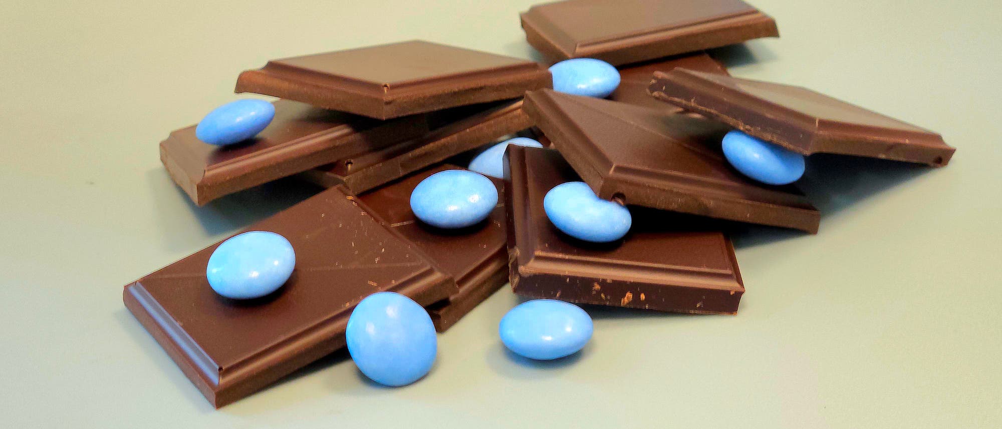 Ein Haufen von Schokoladenst&uuml;cken, durchsetzt mit blauen Schokolinsen, liegt auf einer glatten, hellen Oberfl&auml;che. Die Schokoladenst&uuml;cke sind ungleichm&auml;&szlig;ig gestapelt, w&auml;hrend die blauen Linsen zuf&auml;llig dazwischen verteilt sind. Der Hintergrund ist neutral und lenkt nicht vom Hauptmotiv ab.