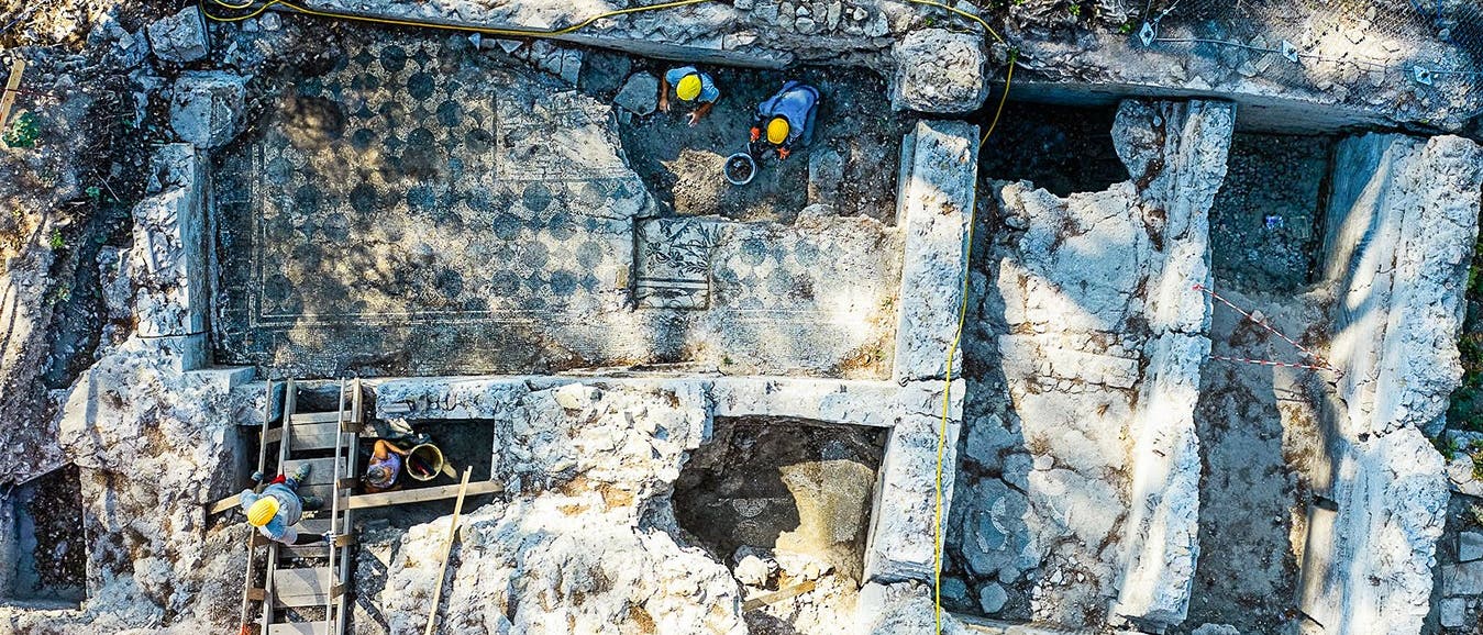 Luftaufnahme einer archäologischen Ausgrabungsstätte mit freigelegten Mauerresten und einem Mosaikboden. Drei Personen mit Schutzhelmen arbeiten an der Stätte, umgeben von Werkzeugen und Ausrüstungen. Die Szene zeigt die Struktur der antiken Ruinen und die laufenden archäologischen Arbeiten. Luftaufnahme einer archäologischen Ausgrabungsstätte mit freigelegten Mauerresten und einem Mosaikboden. Drei Personen mit Schutzhelmen arbeiten an der Stätte, umgeben von Werkzeugen und Ausrüstungen. Die Szene zeigt die Struktur der antiken Ruinen und die laufenden archäologischen Arbeiten.