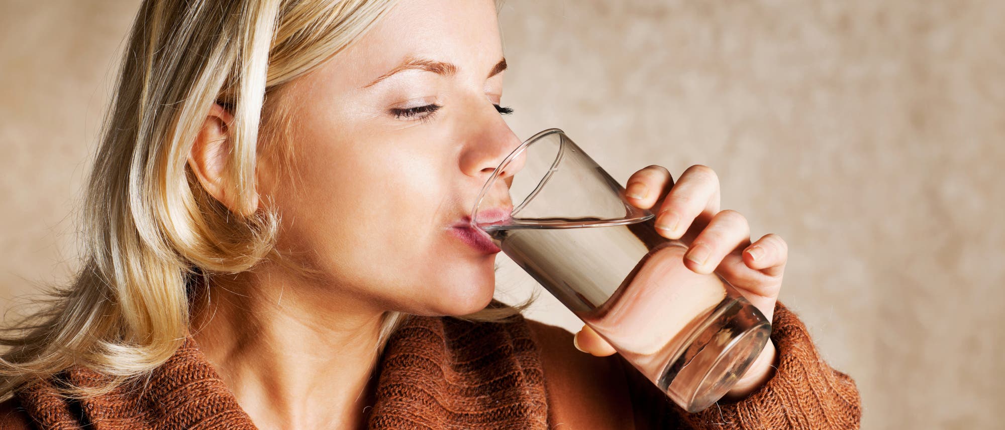 Eine blonde Frau in ockerfarbenen Pullover trinkt aus einem Glas Wasser. 