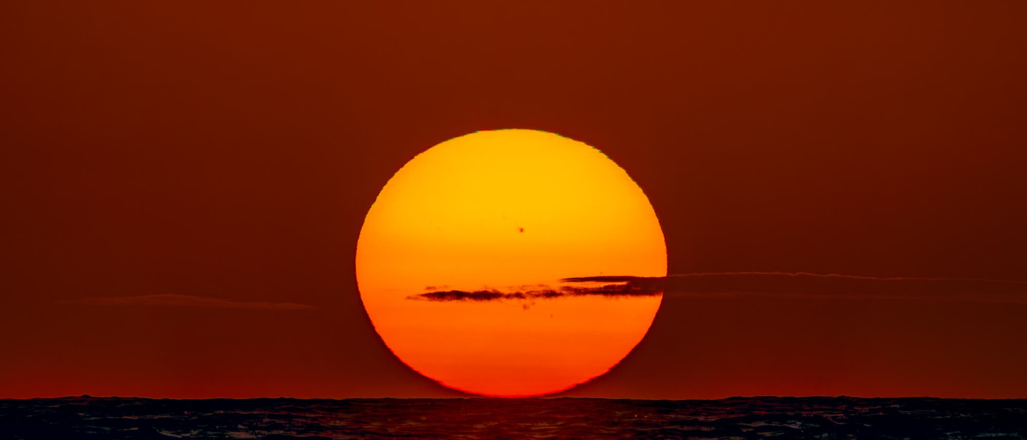 Sonnenuntergang &uuml;ber dem Meer mit einer gro&szlig;en, leuchtend orangefarbenen Sonne am Horizont. Der Himmel ist in warmen Rott&ouml;nen gef&auml;rbt, und die Wasseroberfl&auml;che reflektiert das Licht der untergehenden Sonne. Ein paar Wolken ziehen vor der Sonne vorbei.