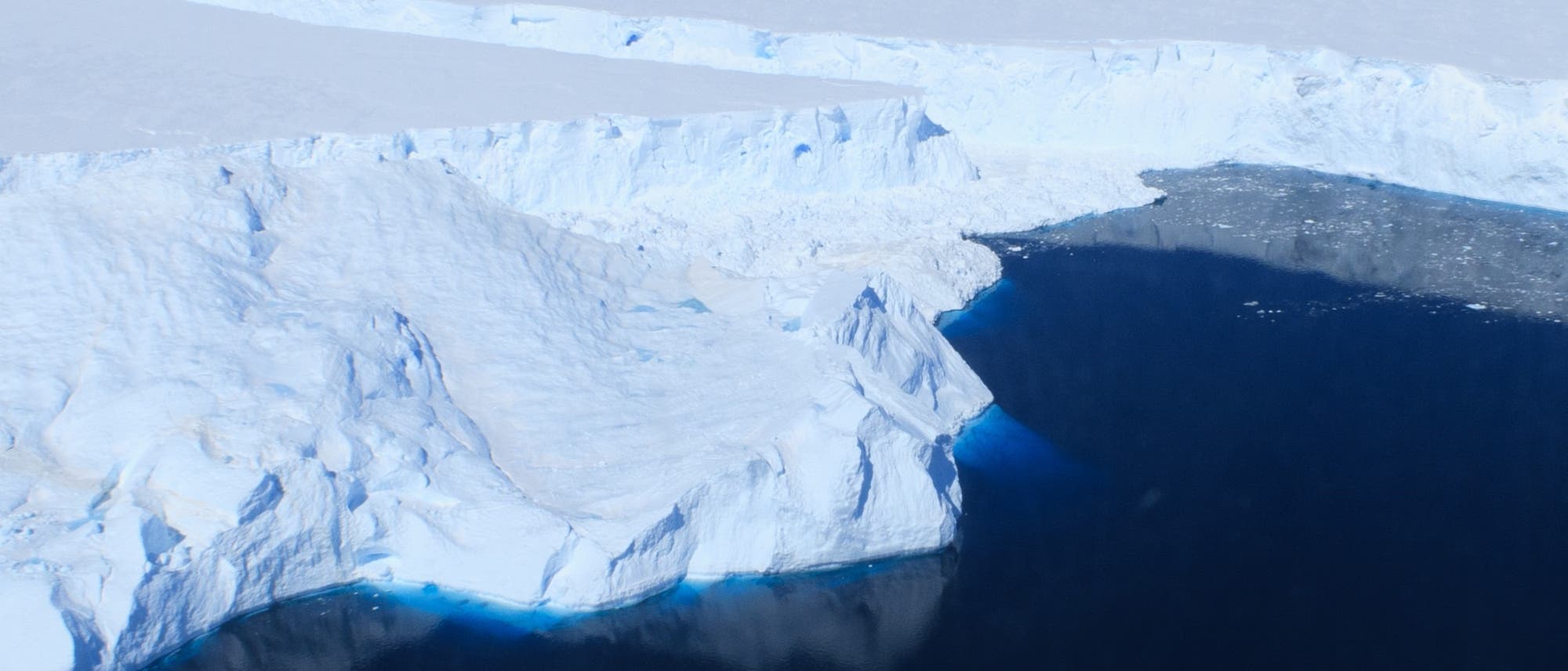 Luftaufnahme eines großen Gletschers, der in ein dunkles Meer mündet. Die weiße Eisfläche erstreckt sich weit in den Hintergrund, während das Wasser im Vordergrund dunkelblau erscheint. Der Himmel ist klar und blau, was die Weite und Kälte der Landschaft betont. Luftaufnahme eines großen Gletschers, der in ein dunkles Meer mündet. Die weiße Eisfläche erstreckt sich weit in den Hintergrund, während das Wasser im Vordergrund dunkelblau erscheint. Der Himmel ist klar und blau, was die Weite und Kälte der Landschaft betont.