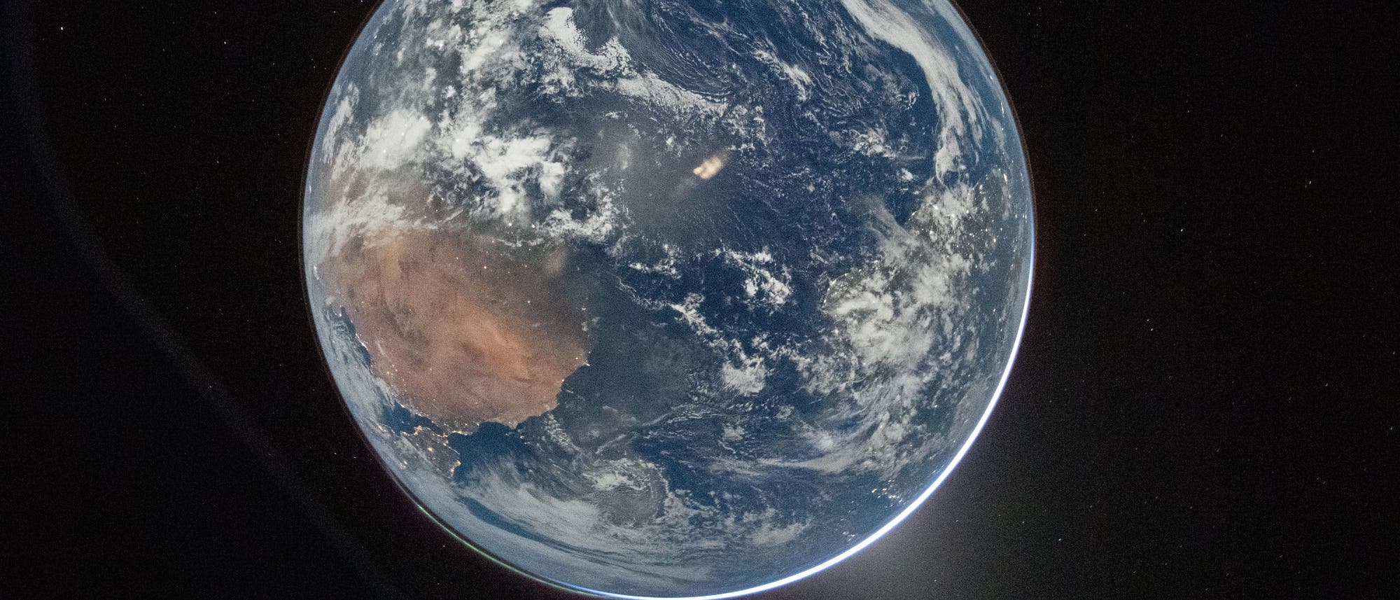 Ansicht der Erde aus dem Weltraum, die den Planeten in seiner vollen Kugelform zeigt. Die Oberfl&auml;che ist mit Wolken bedeckt, und Kontinente sind sichtbar, umgeben von Ozeanen. Der Weltraum im Hintergrund ist dunkel mit einigen sichtbaren Sternen. Die Kr&uuml;mmung der Erde ist deutlich erkennbar, und ein heller Lichtschein umgibt den Planeten.