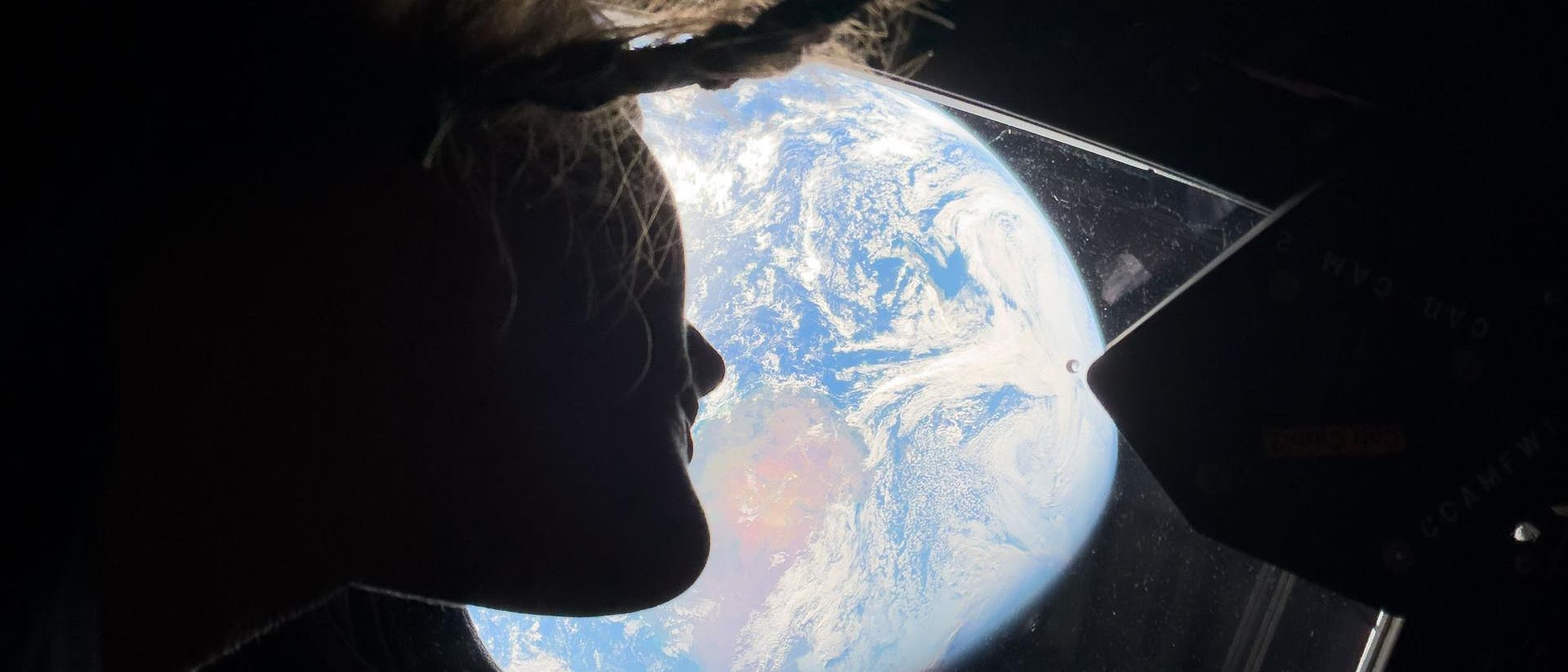 Vista de la Tierra desde la ventana de una nave espacial. Una persona mira por la ventana mientras la Tierra es visible en todo su esplendor con océanos azules y formaciones de nubes blancas. La escena transmite la impresión de ingravidez y de inmensidad del espacio.