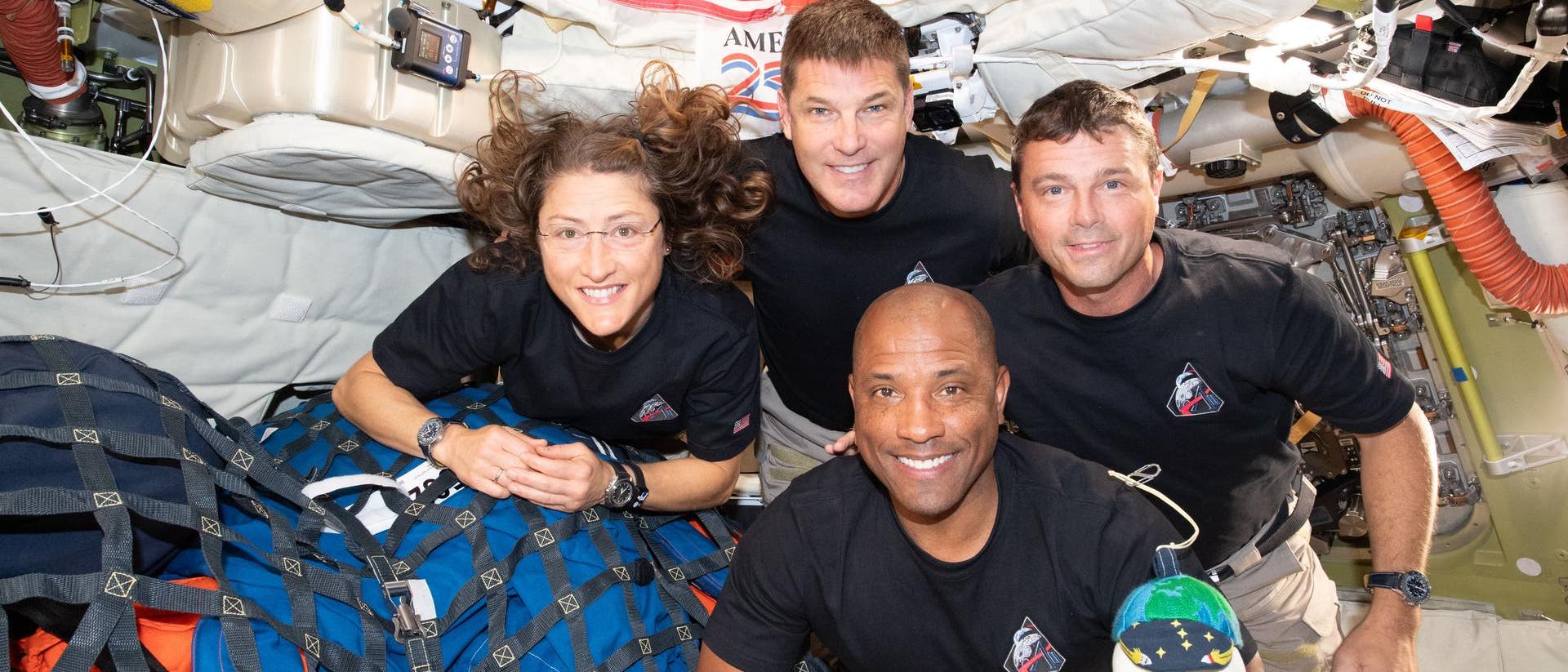 Eine Gruppe von vier Personen in einem Raumfahrzeug, umgeben von technischen Geräten und Kabeln. Sie tragen schwarze T-Shirts mit einem Logo und lächeln in die Kamera. Im Hintergrund sind die Flaggen der USA und Kanadas sichtbar. Die Umgebung wirkt beengt und funktional, typisch für eine Raumstation. Eine Gruppe von vier Personen in einem Raumfahrzeug, umgeben von technischen Geräten und Kabeln. Sie tragen schwarze T-Shirts mit einem Logo und lächeln in die Kamera. Im Hintergrund sind die Flaggen der USA und Kanadas sichtbar. Die Umgebung wirkt beengt und funktional, typisch für eine Raumstation.