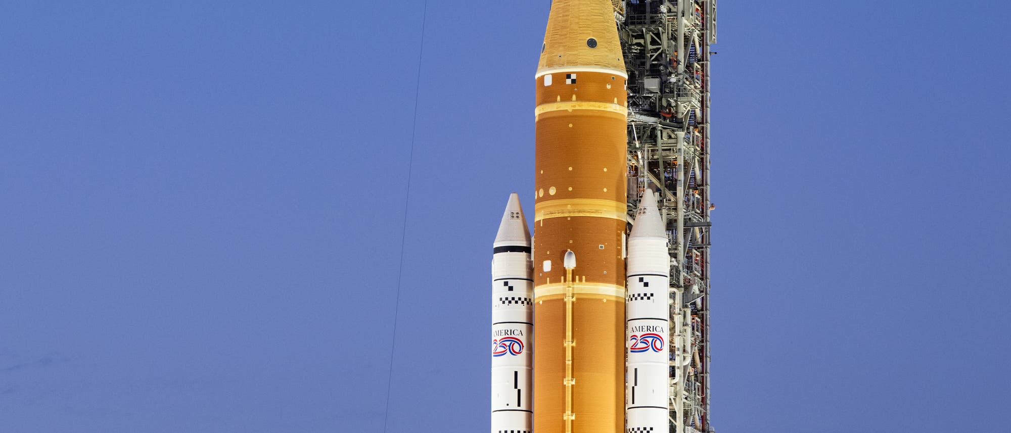 Eine gro&szlig;e Rakete steht auf einer Startrampe vor einem klaren, blauen Himmel. Die Rakete ist mit dem Logo einer bekannten Raumfahrtbeh&ouml;rde und der Aufschrift "USA" versehen. Im Hintergrund ist eine komplexe Struktur aus Metallger&uuml;sten zu sehen. Am unteren Rand des Bildes ist eine gro&szlig;e amerikanische Flagge angebracht. Die Szene ist gut beleuchtet, was auf eine bevorstehende Mission hindeutet.