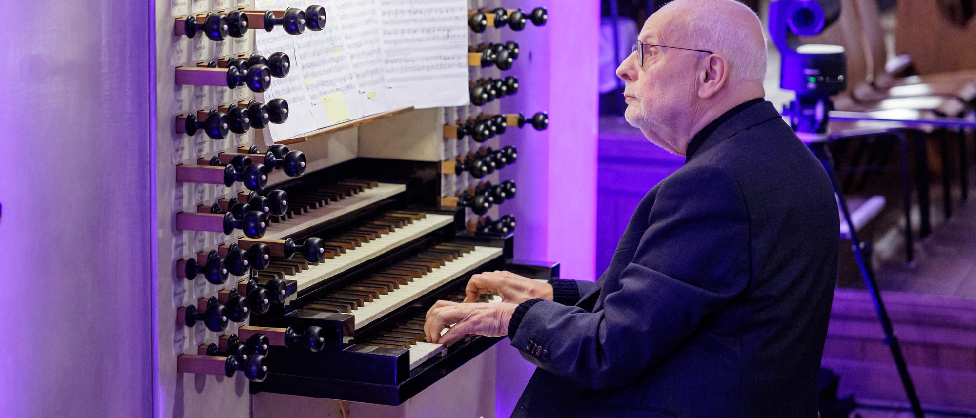 Ein älterer Mann spielt konzentriert auf einer großen Kirchenorgel. Die Orgel hat mehrere Manuale und zahlreiche Registerzüge. Notenblätter sind auf dem Notenständer platziert. Der Raum ist in violettes Licht getaucht, und im Hintergrund sind Kirchenbänke zu sehen. Ein älterer Mann spielt konzentriert auf einer großen Kirchenorgel. Die Orgel hat mehrere Manuale und zahlreiche Registerzüge. Notenblätter sind auf dem Notenständer platziert. Der Raum ist in violettes Licht getaucht, und im Hintergrund sind Kirchenbänke zu sehen.