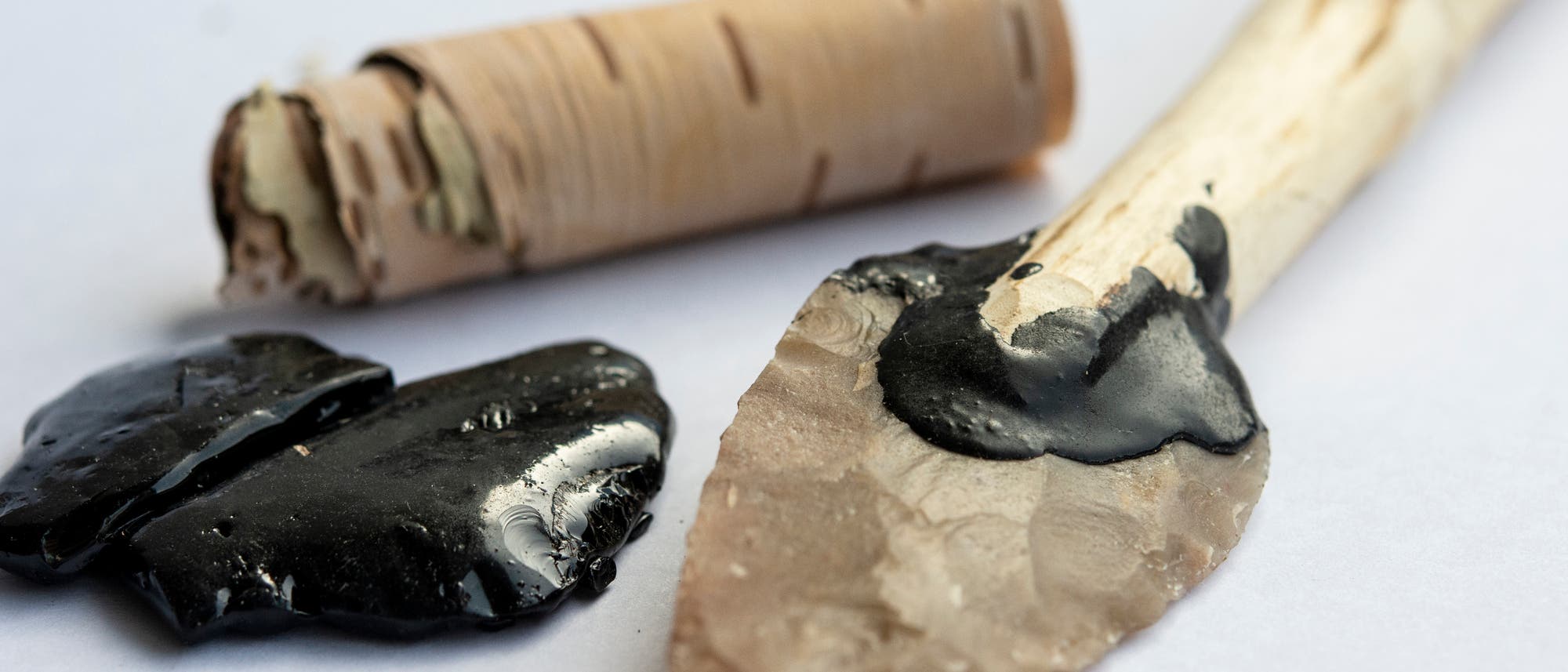 Ein prähistorischer Speer mit einer Spitze aus geschliffenem Stein, befestigt an einem Holzschaft mit schwarzem Birkenpech. Neben dem Speer liegt ein Stück Birkenrinde und zwei Kleckse Birkenpech auf einer weißen Oberfläche.