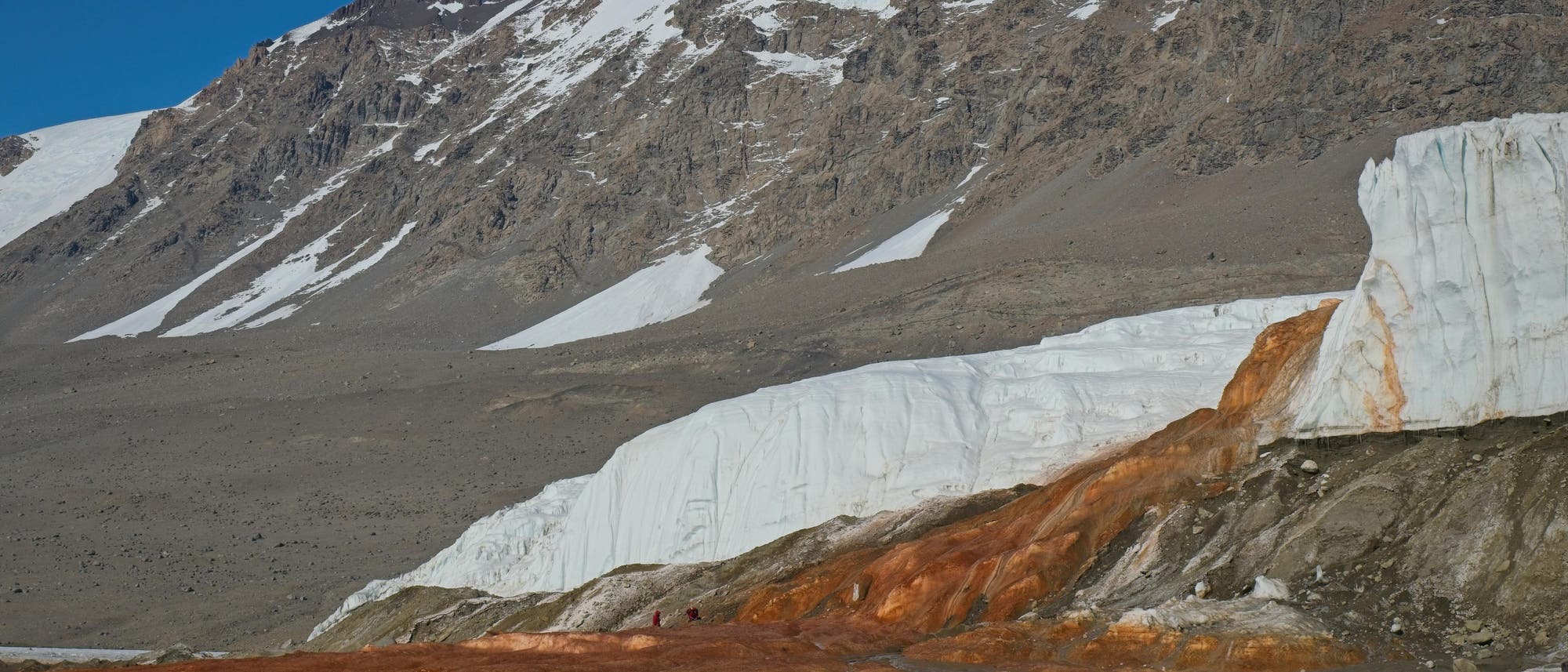 Eine Landschaftsaufnahme zeigt eine felsige Bergkette mit schneebedeckten Gipfeln unter einem klaren blauen Himmel. Im Vordergrund ist ein Gletscher zu sehen, aus dem rostfarbenes Wasser fließt, das sich über das Eis und den Boden erstreckt. Die Szene vermittelt eine karge, aber beeindruckende natürliche Umgebung. Eine Landschaftsaufnahme zeigt eine felsige Bergkette mit schneebedeckten Gipfeln unter einem klaren blauen Himmel. Im Vordergrund ist ein Gletscher zu sehen, aus dem rostfarbenes Wasser fließt, das sich über das Eis und den Boden erstreckt. Die Szene vermittelt eine karge, aber beeindruckende natürliche Umgebung.