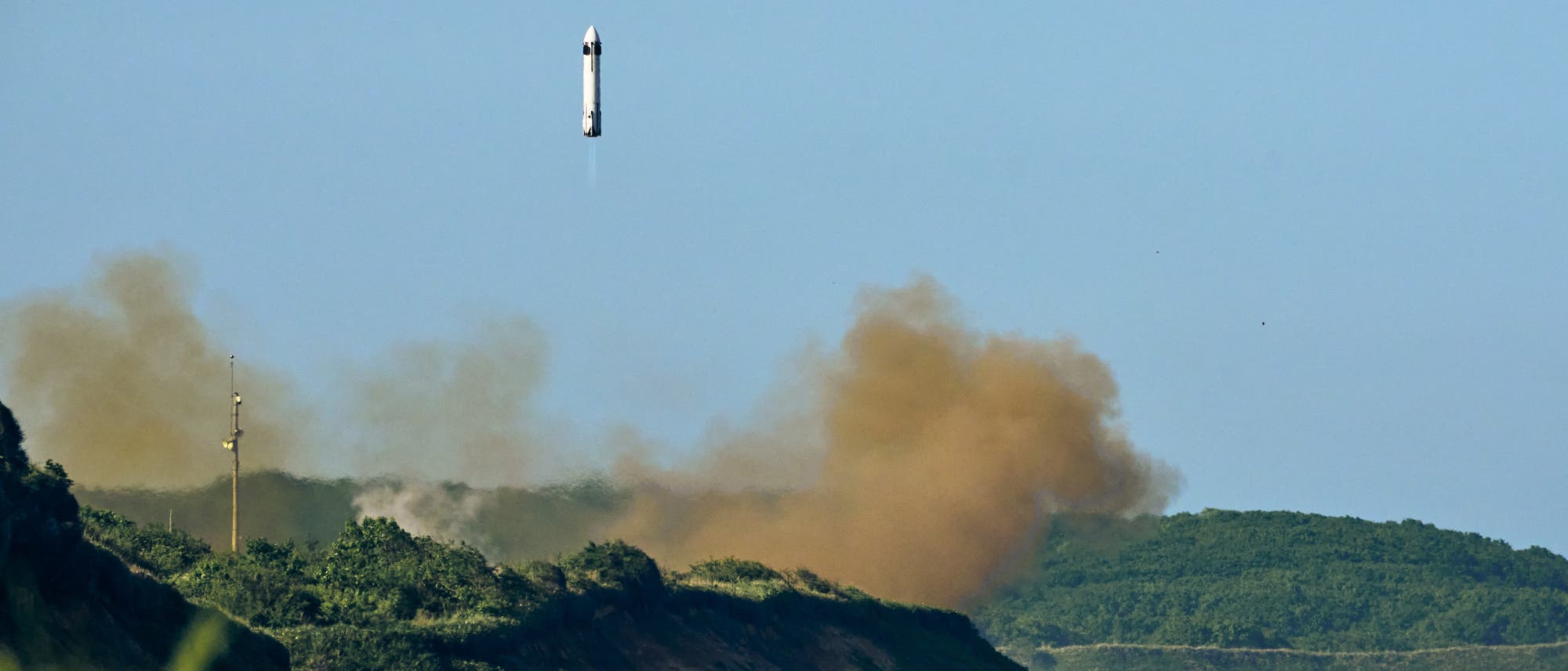 Eine Rakete steigt senkrecht in den blauen Himmel auf, während sie von einer grünen, bewaldeten Landschaft umgeben ist. Am Boden ist eine große Staubwolke zu sehen, die den Start begleitet. Im Hintergrund erstrecken sich Hügel.