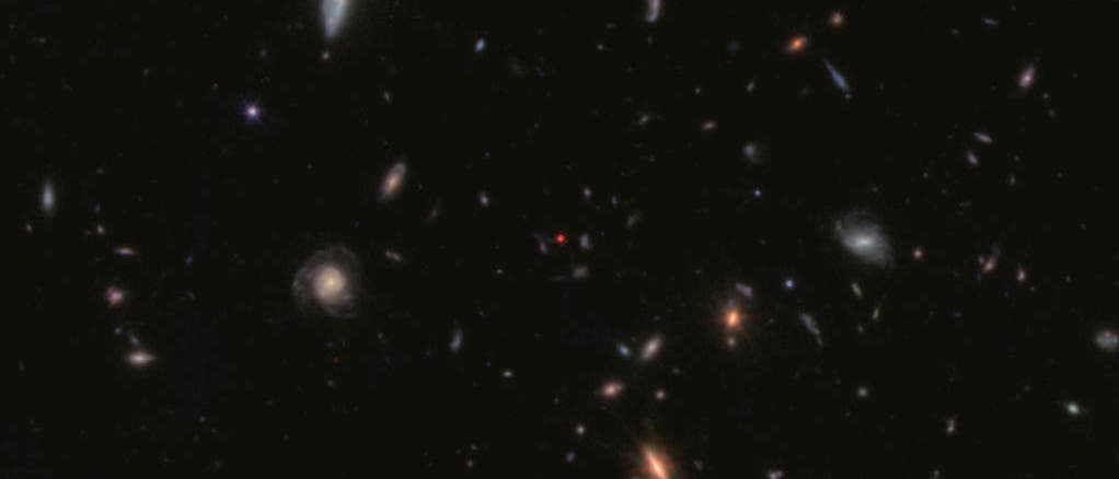 Die Aufnahme zeigt mehrere leuchtende Galaxien mit unterschiedlichen Formen. In der Mitte befindet sich ein rot markierter Punkt. 