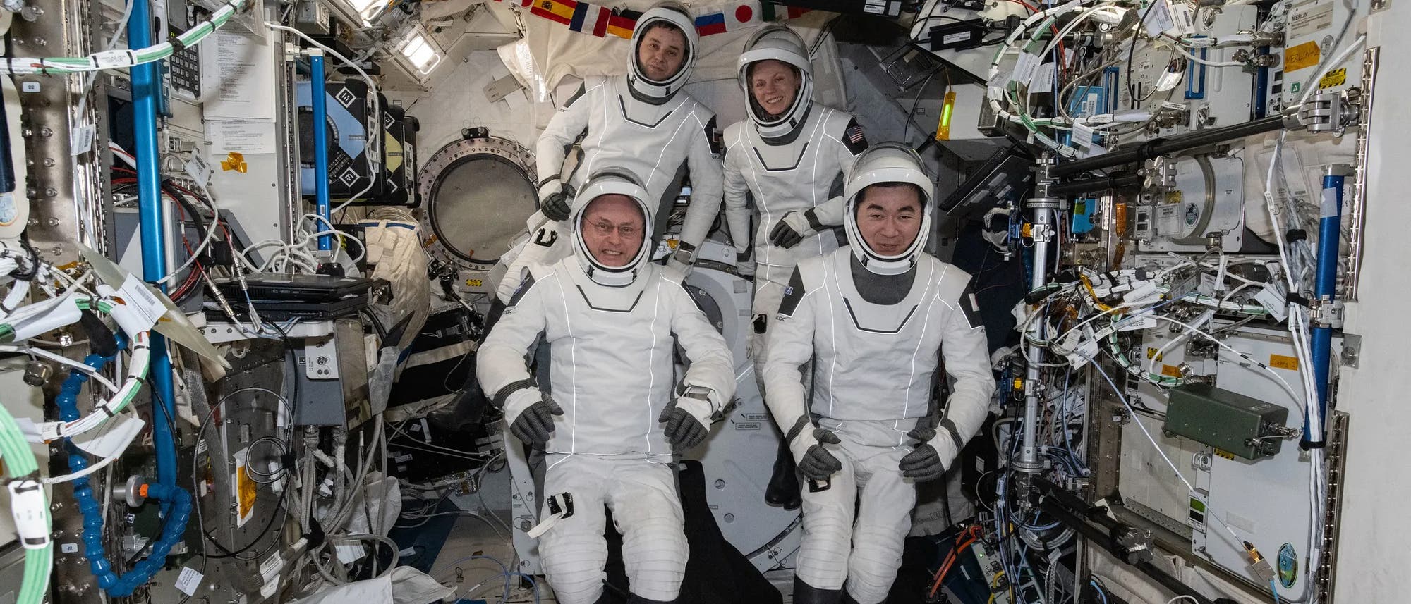 Eine Gruppe von vier Astronauten in wei&szlig;en Raumanz&uuml;gen posiert in einem beengten, technisch ausgestatteten Raumfahrzeugmodul. Die Umgebung ist mit zahlreichen Kabeln, Ger&auml;ten und Kontrolltafeln ausgestattet. Im Hintergrund sind Flaggen verschiedener L&auml;nder zu sehen. Die Astronauten l&auml;cheln und blicken in die Kamera, was auf eine positive Stimmung hinweist.
