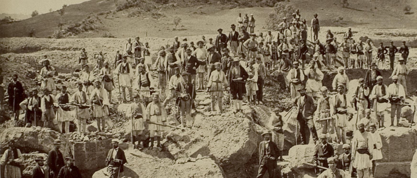 Eine historische Fotografie zeigt eine gro&szlig;e Gruppe von Menschen, die auf und um gro&szlig;e Felsbl&ouml;cke und griechische Ruinen herumstehen. Viele Personen tragen traditionelle Kleidung und posieren f&uuml;r das Foto. Im Hintergrund ist eine h&uuml;gelige Landschaft zu sehen. 